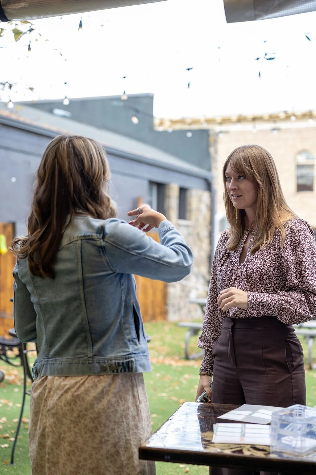 Two women having a conversation outdoors, one gesturing with her hand, in a backyard with string lights and picnic tables.