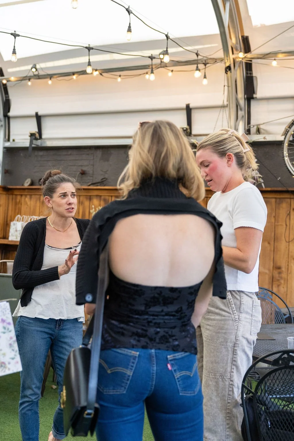Three women engaged in conversation inside a cozy venue with string lights overhead. One woman with a bun is gesturing with her hand, another with blonde hair and a pink bow is looking down, and the third has her back to the camera showing a black to