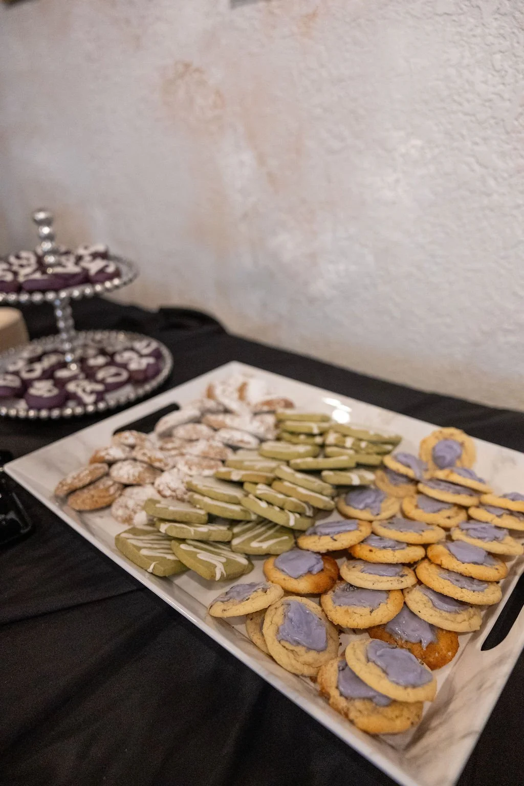 Plate of assorted cookies with purple frosting, piped cookies with green and white icing, and sugar cookies with powdered sugar, on a black table with a three-tiered purple and silver dessert stand in the background.
