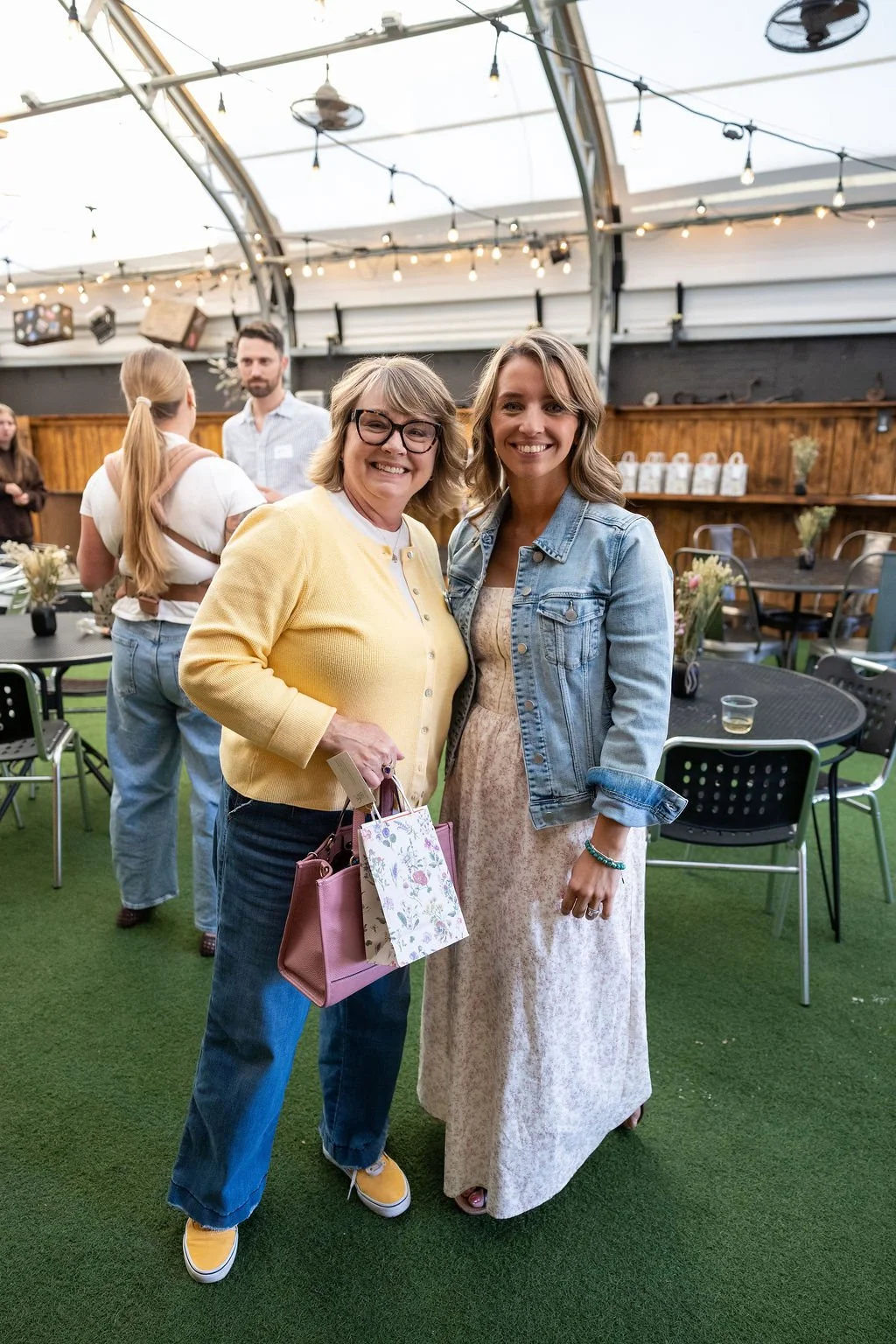 Two women standing together and smiling at an indoor event, with other guests and tables in the background.