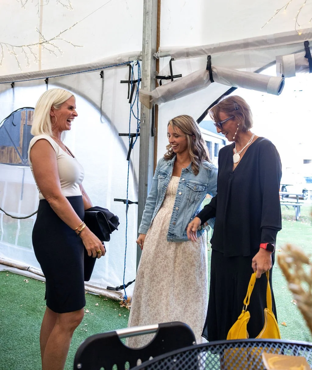 Three women are laughing and engaging in conversation at an outdoor gathering under a tent with string lights, with grass on the ground and a building in the background.