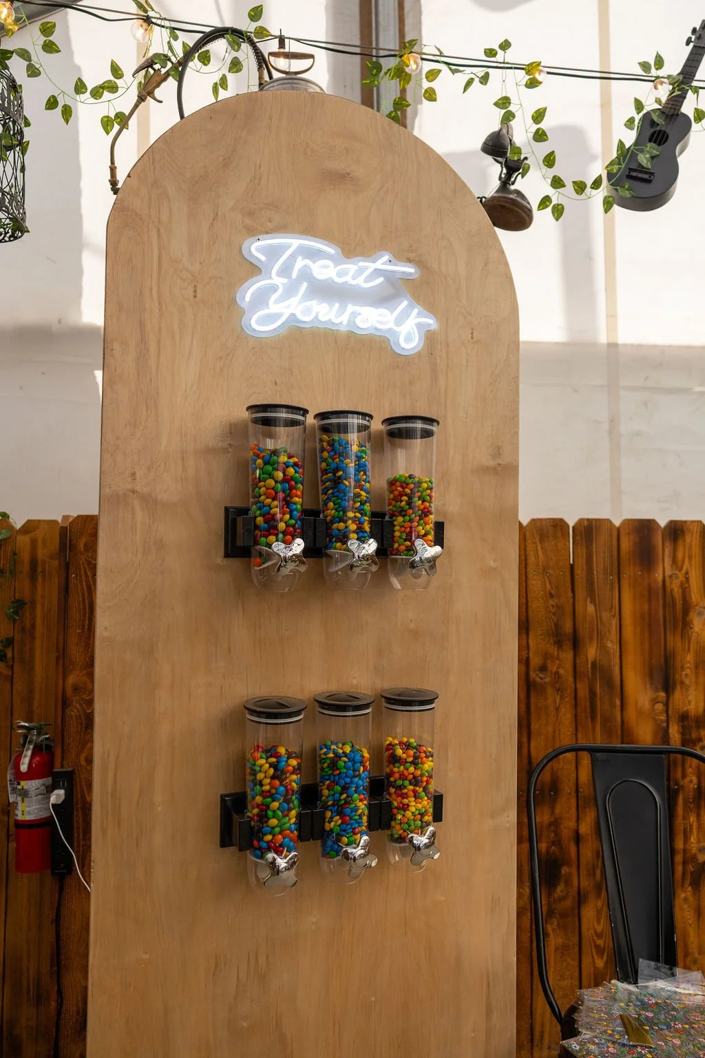 A gumball machine display with six transparent containers filled with colorful gumballs, three on the top row and three on the bottom, with a neon sign reading 'Treat Yourself' above.