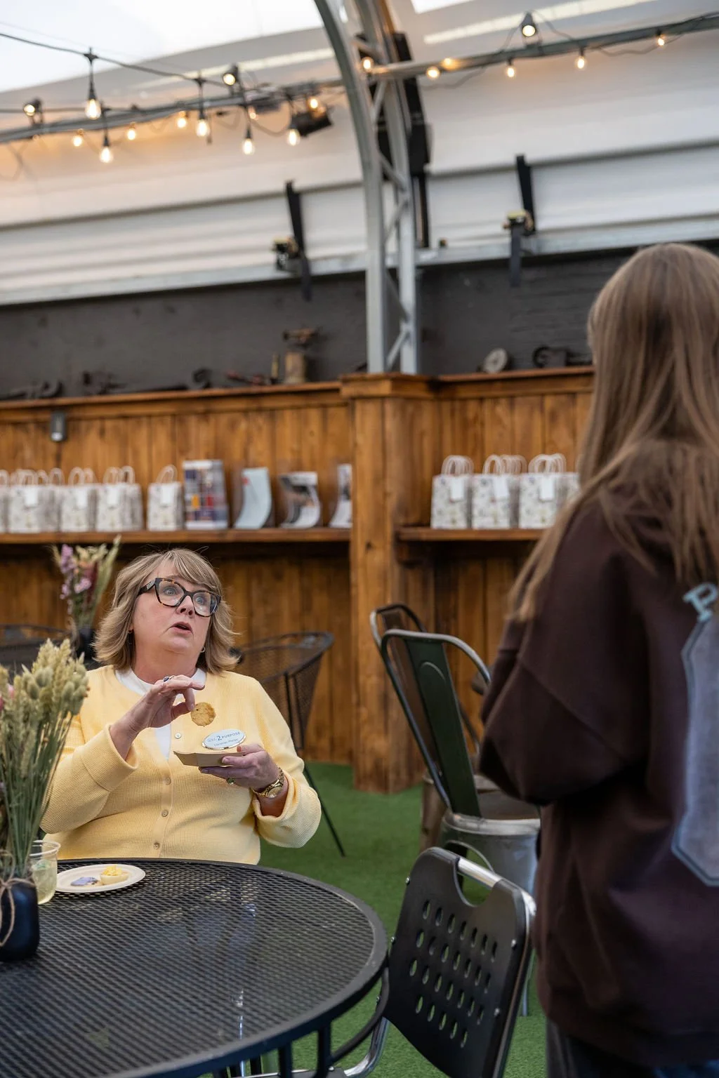 A woman with glasses and a yellow sweater sitting at a black metal outdoor dining table, holding a plate and a snack, talking to another woman in a brown jacket at a social gathering under a covered outdoor area with string lights and wooden walls.