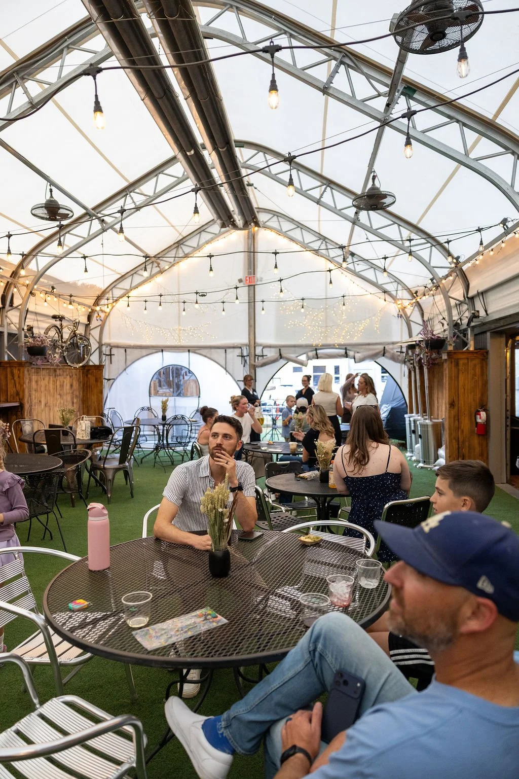 People sitting and standing at tables inside a decorated, semi-outdoor event tent with string lights, fans, and bikes hanging from the ceiling