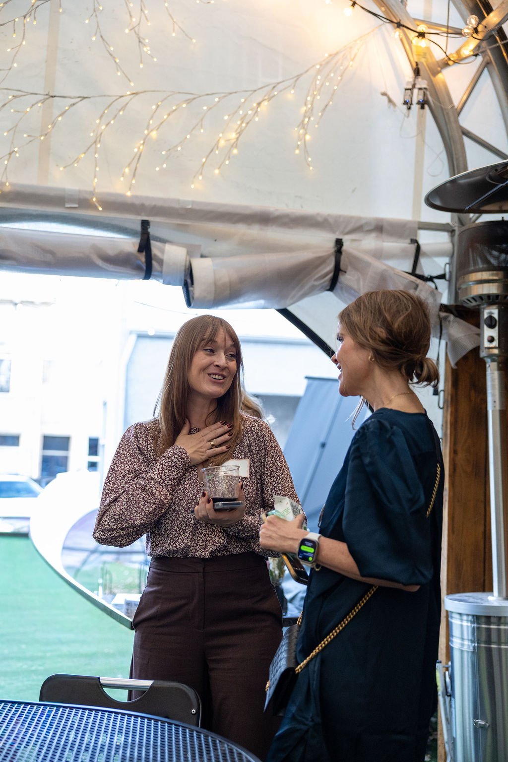 Two women are talking and smiling inside a tent with decorative string lights hanging above them. One woman holds a drink, while the other has a small object and wears a smartwatch. They are near outdoor tables and windows showing a building and park