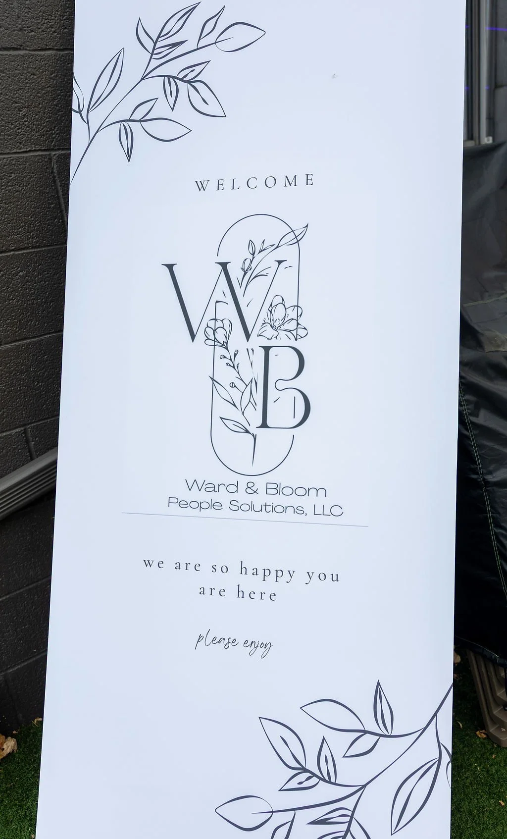 A vertical sign with a white background and black decorative floral illustrations at the top and bottom. The sign reads 'WELCOME' at the top, with a large stylized 'W' and 'B' in the center, decorated with floral designs. Below, it says "Ward & Bloom