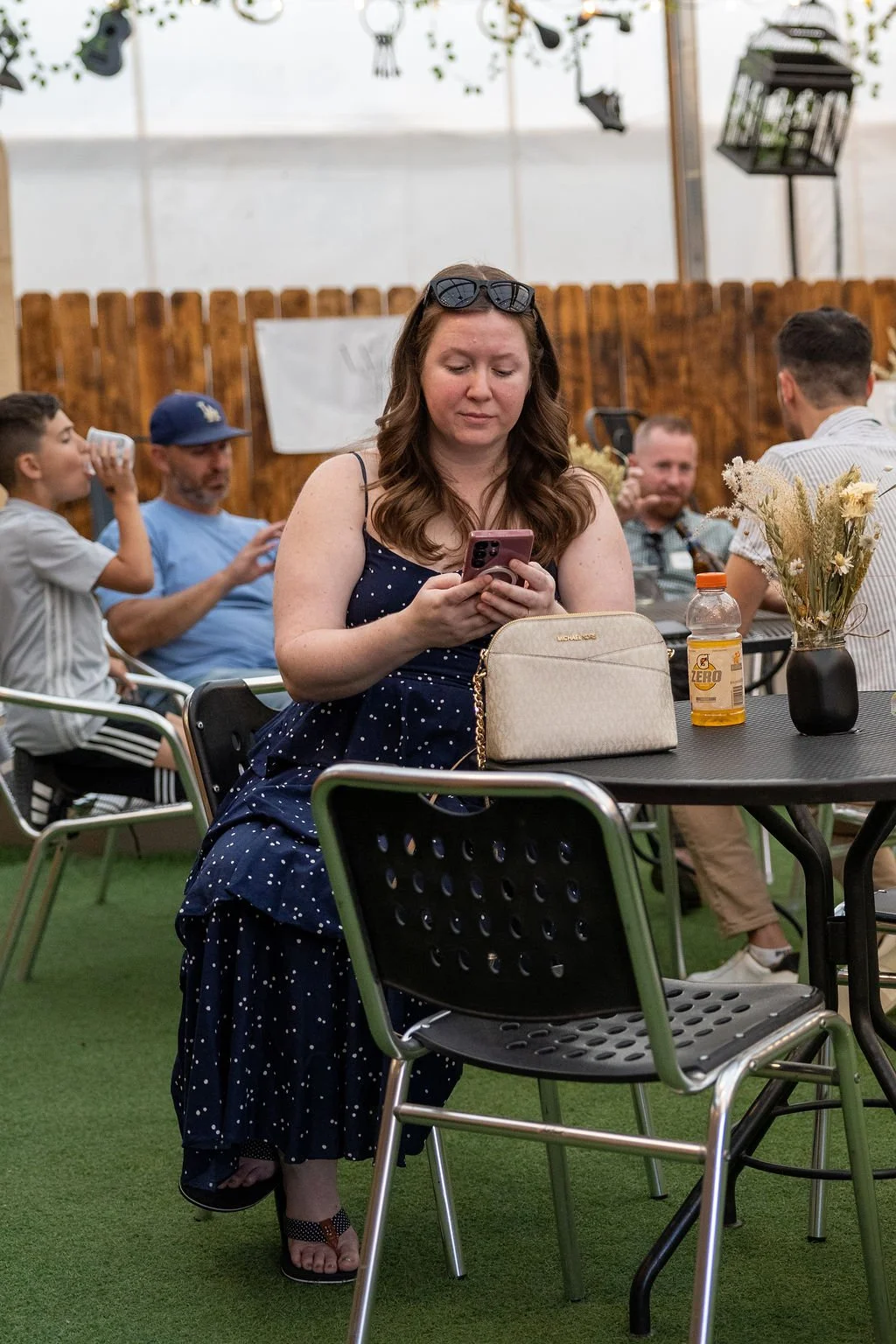 A woman in a navy blue polka dot dress sitting at a black outdoor table, looking at her phone. Glasses rest on her head. On the table are a beige purse, a bottle of Gatorade Zero, and a black vase with dried flowers. In the background, people are sit