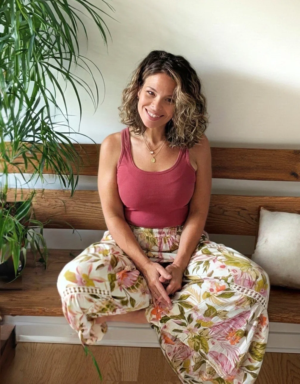 A woman with curly brown hair and a warm smile sitting on a wooden bench, wearing a pink sleeveless top and floral wide-leg pants, next to a large green plant and a white pillow.