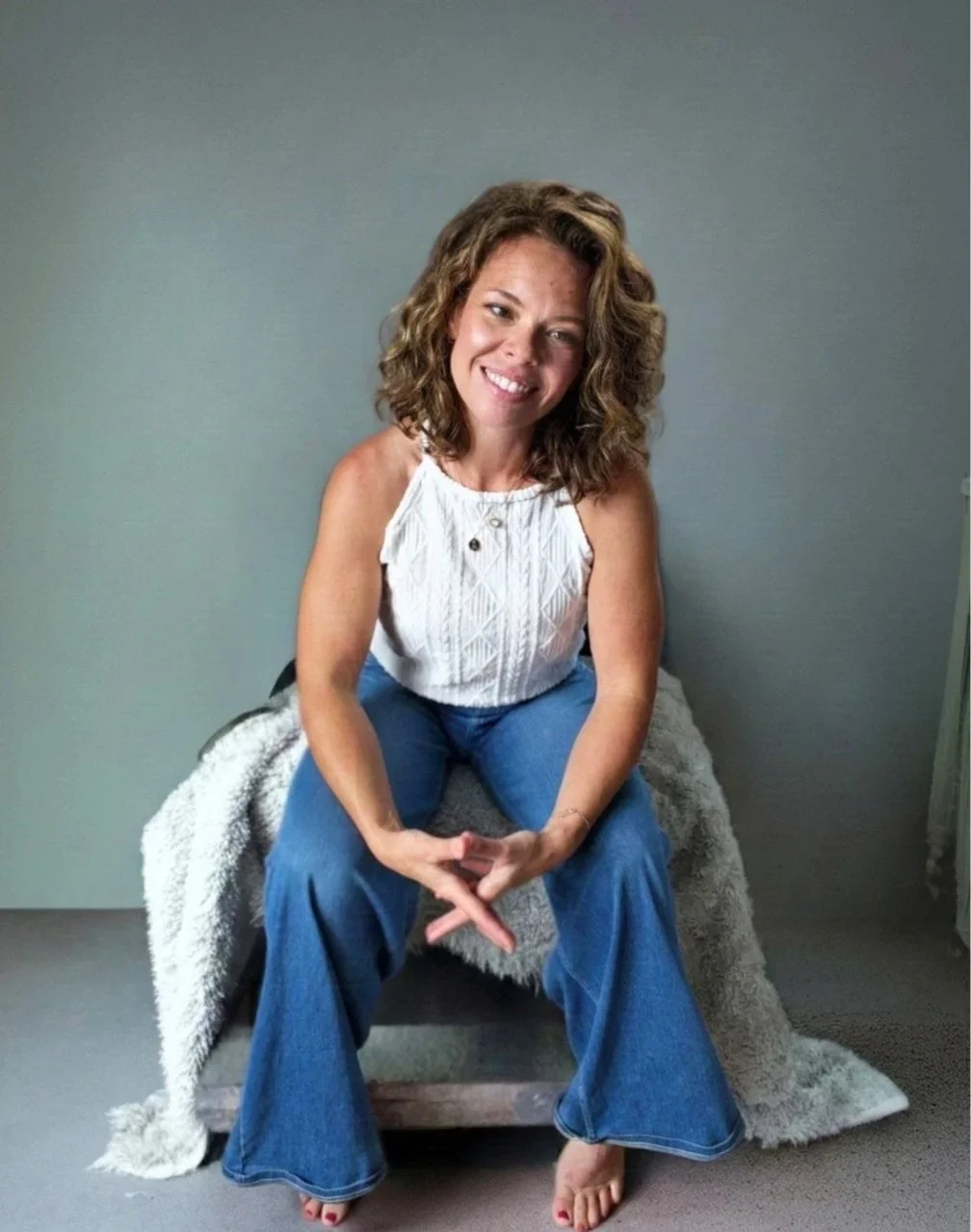 A woman with curly hair sitting on a wooden stool with a grey blanket, smiling at the camera against a plain grey wall.