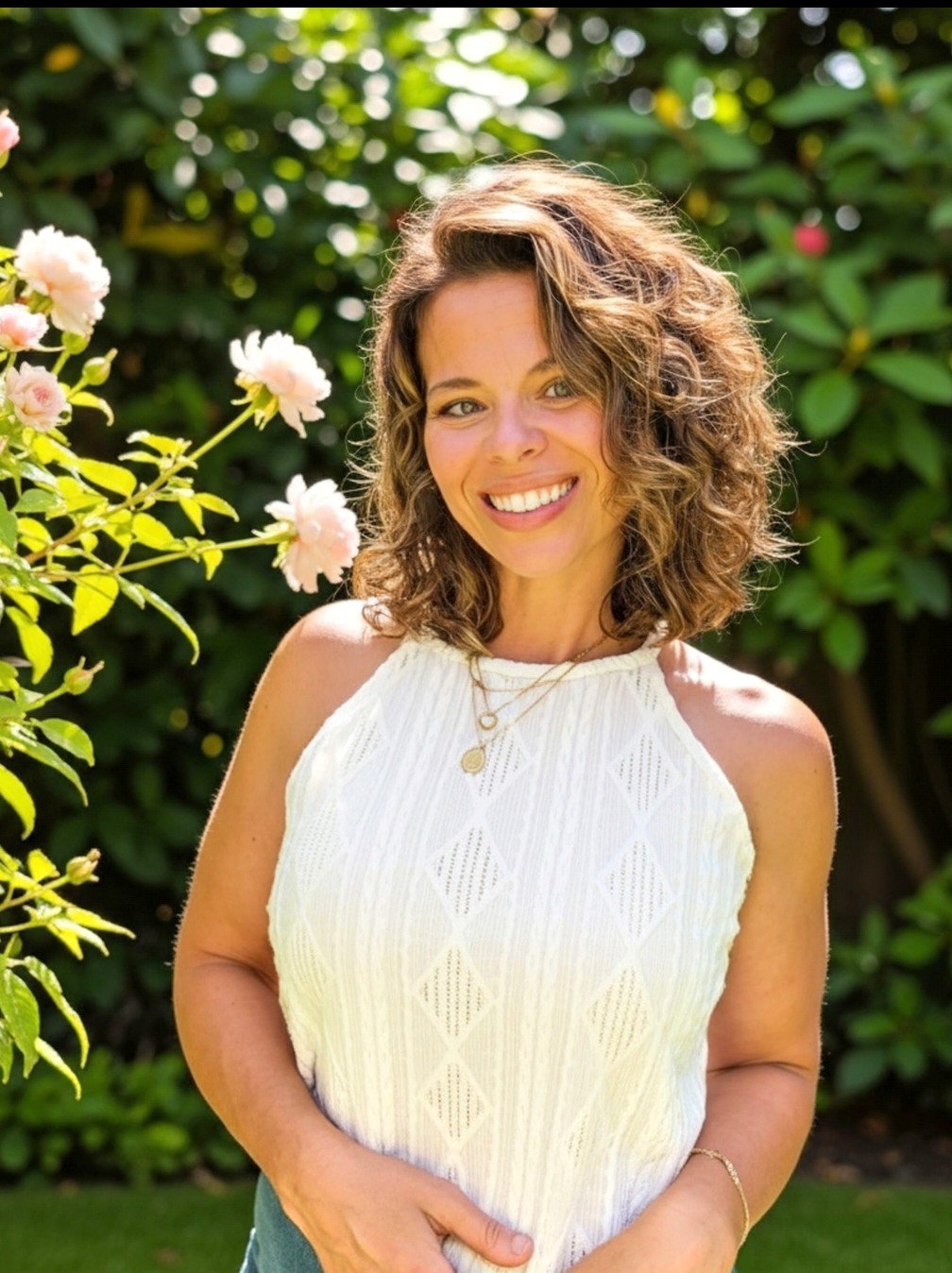 A woman with curly brown hair standing outdoors near pink roses and green foliage, smiling at the camera, wearing a white sleeveless top and layered necklaces.
