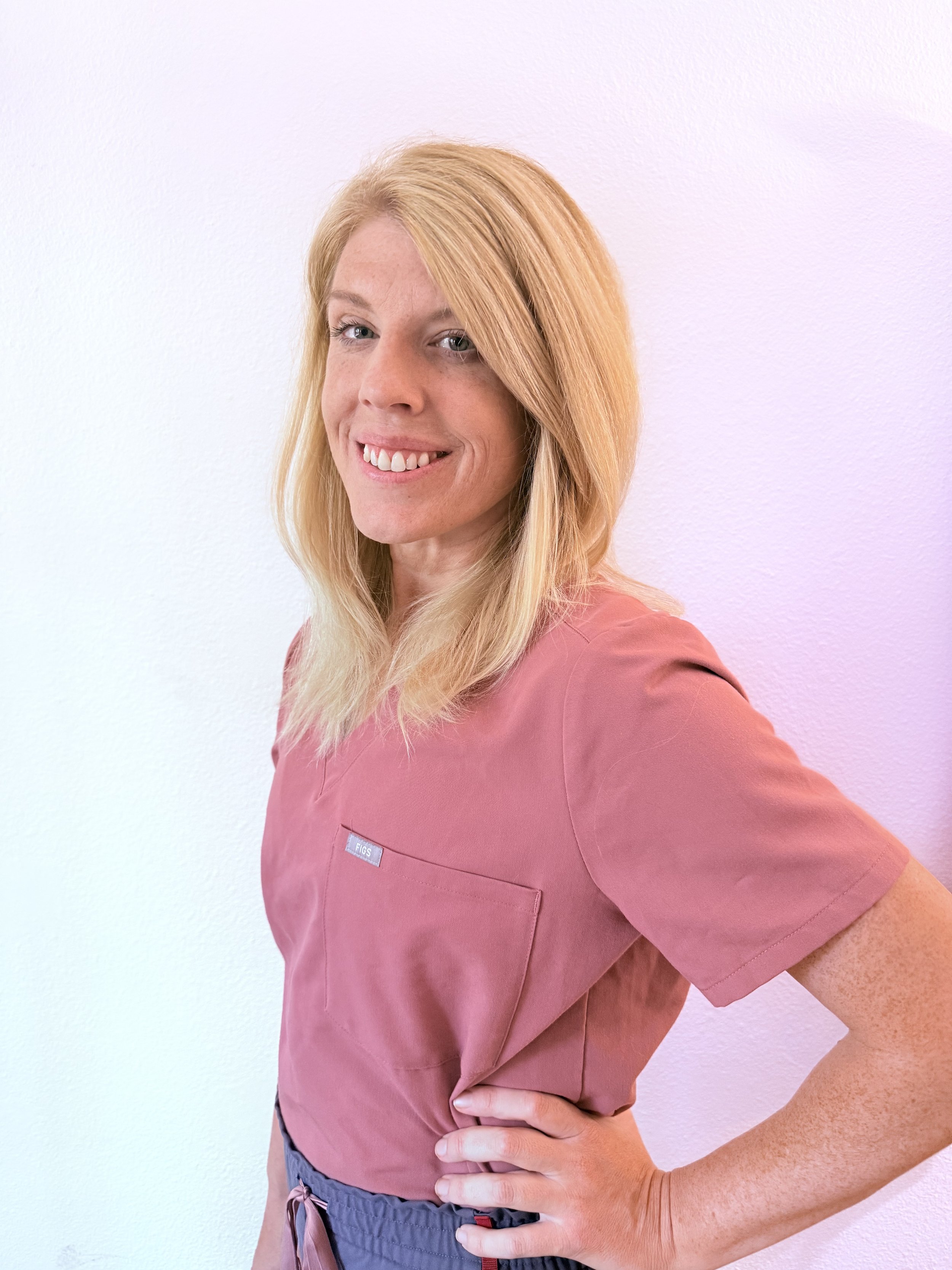A woman with blonde hair in scrubs, standing against a plain white wall, smiling and looking at the camera.