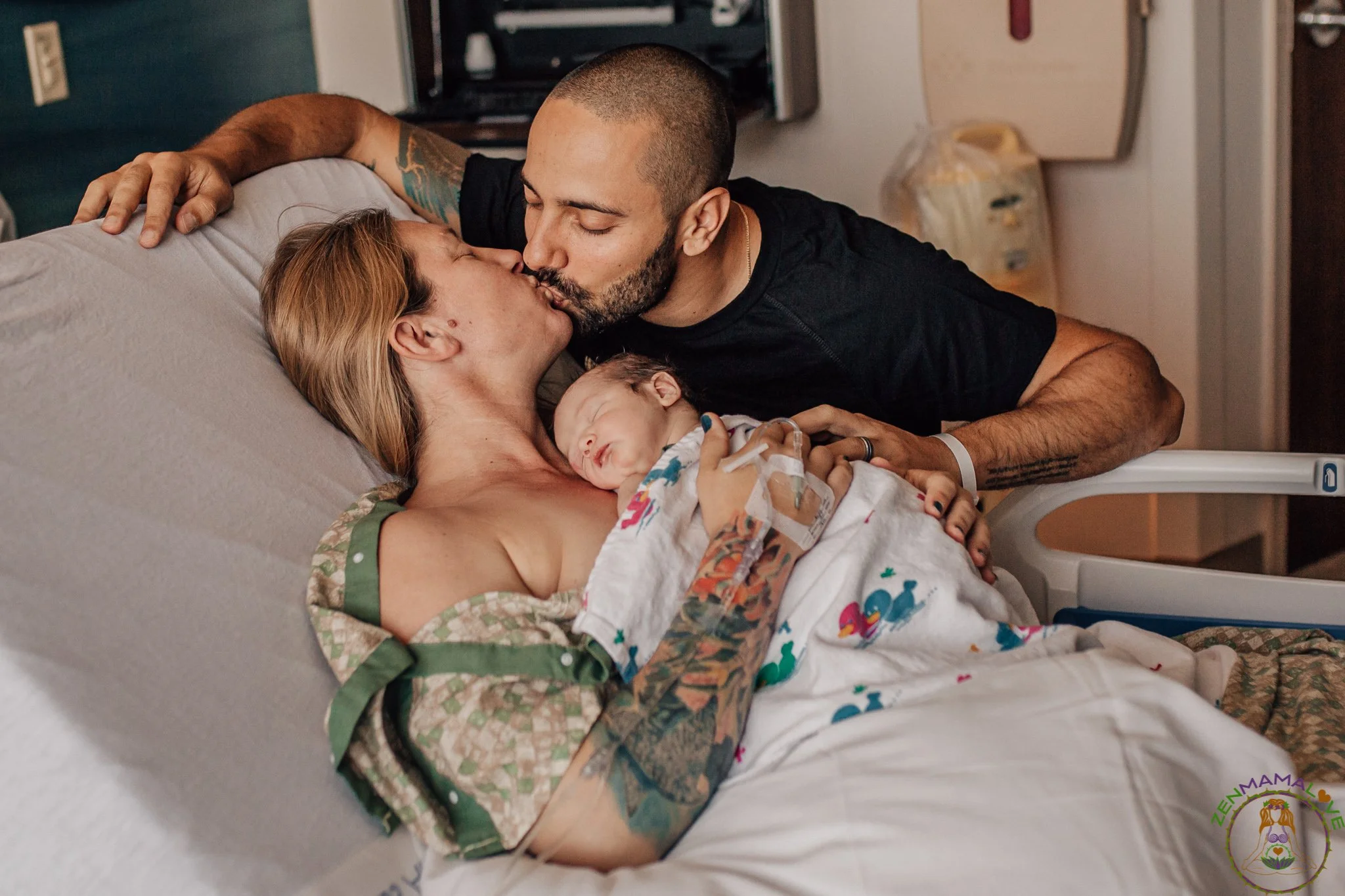 A man with a shaved head and tattoos on his arms kissing a woman with long blonde hair and a tattoo on her neck. They are in a hospital room, lying on a bed with a newborn baby wrapped in a colorful blanket, who is sleeping. The man has a hospital br
