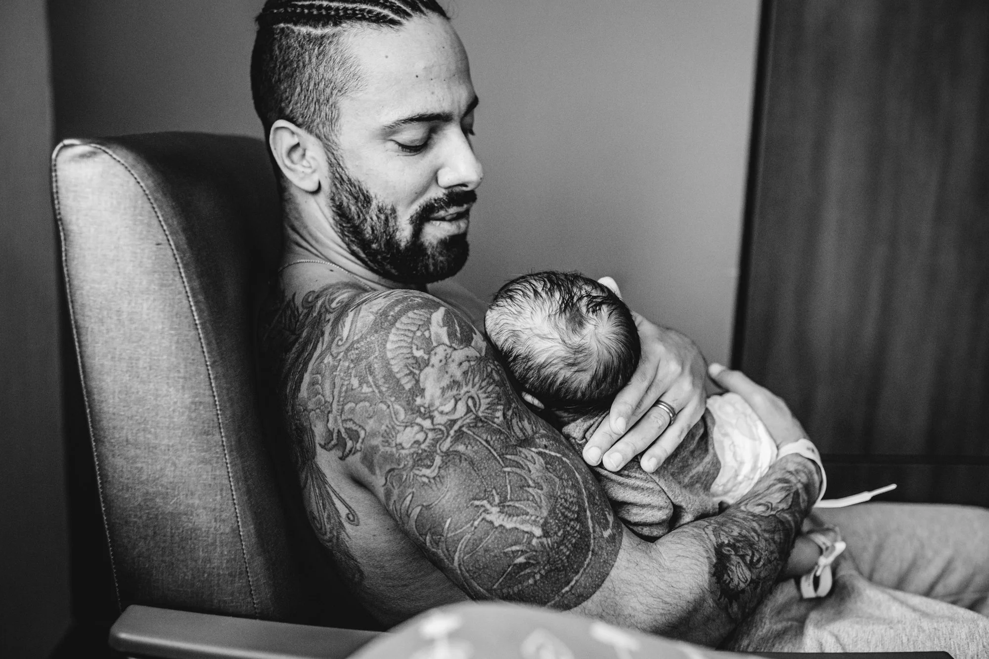 A man with tattoos holding a newborn baby in a hospital room.