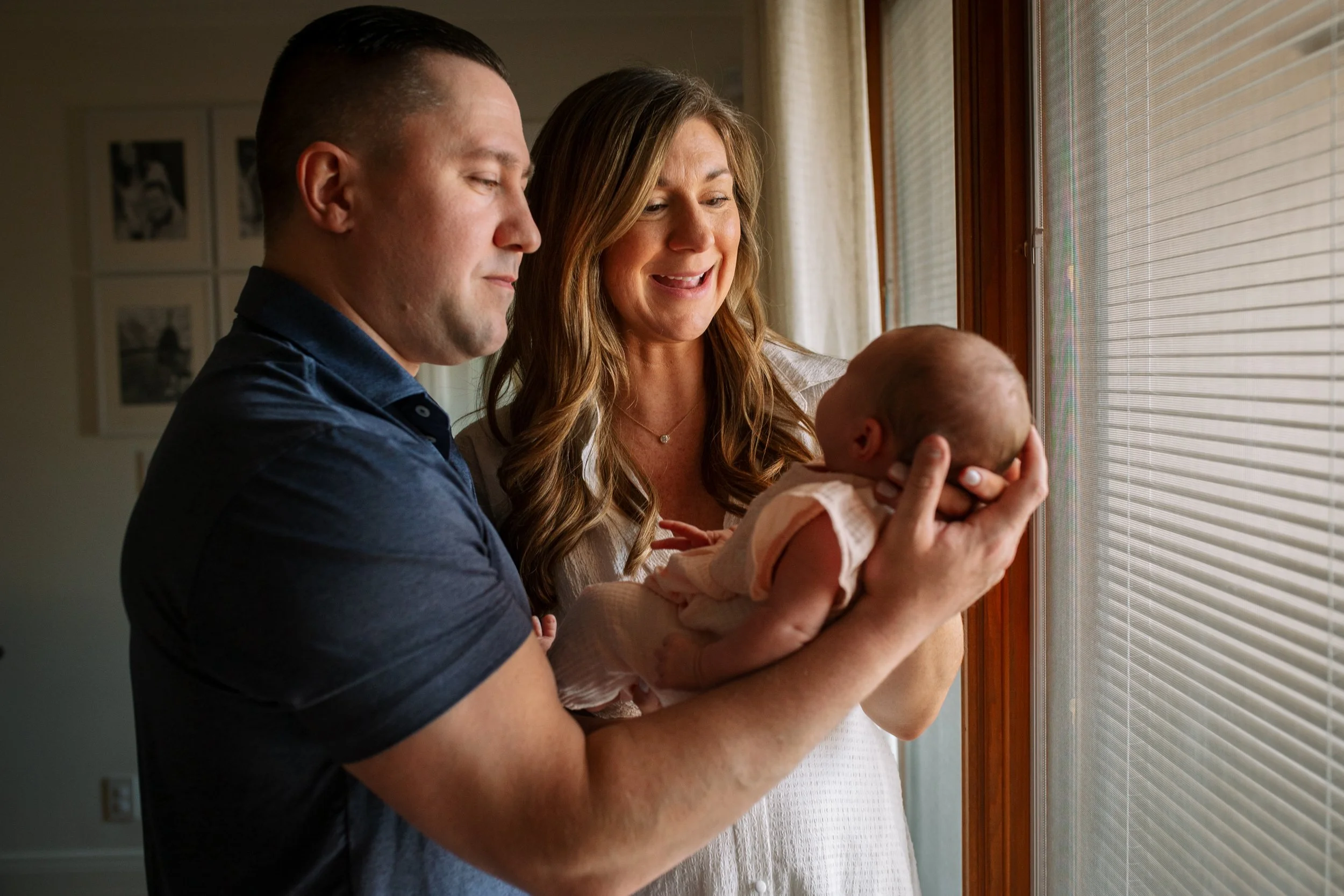 A man and woman holding a newborn baby, looking out a window with blinds in a warmly lit home.