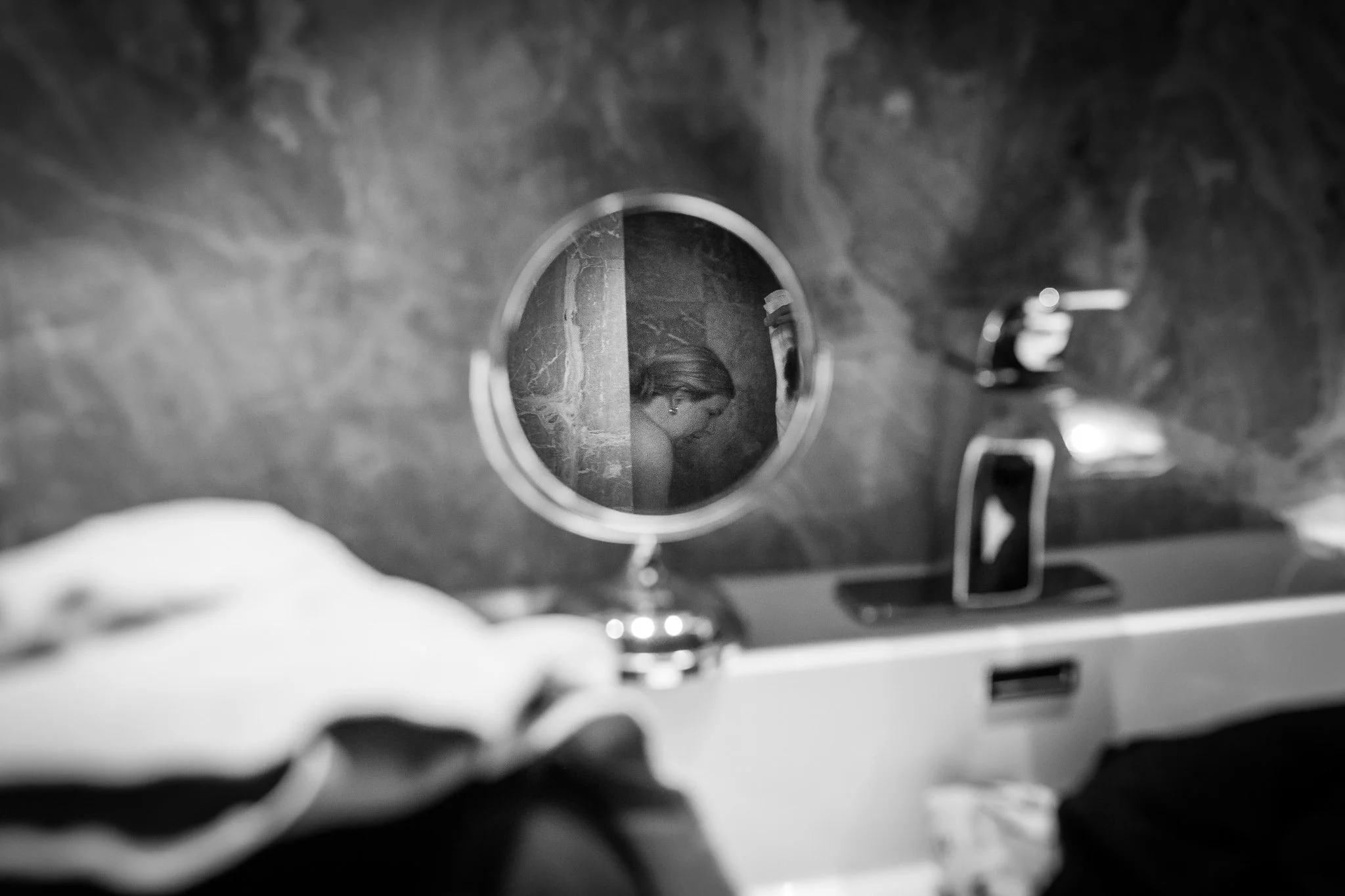 A black and white photo taken through a toilet bowl, showing a woman with long hair looking down. The scene is seen from above, with the toilet bowl's rim framing the woman.