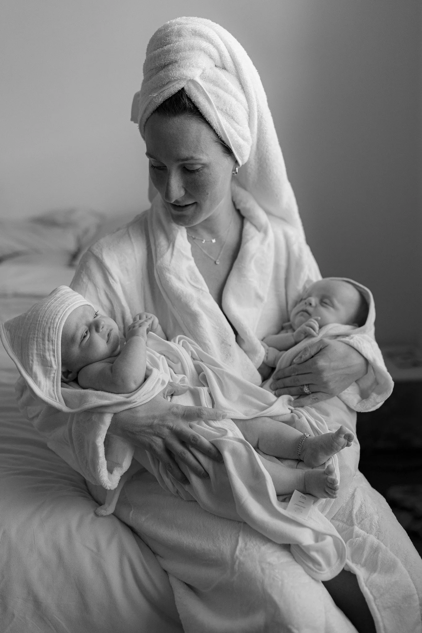 A woman with a towel wrapped around her hair sitting on a bed, holding two newborn babies, one in each arm, in a cozy indoor setting.
