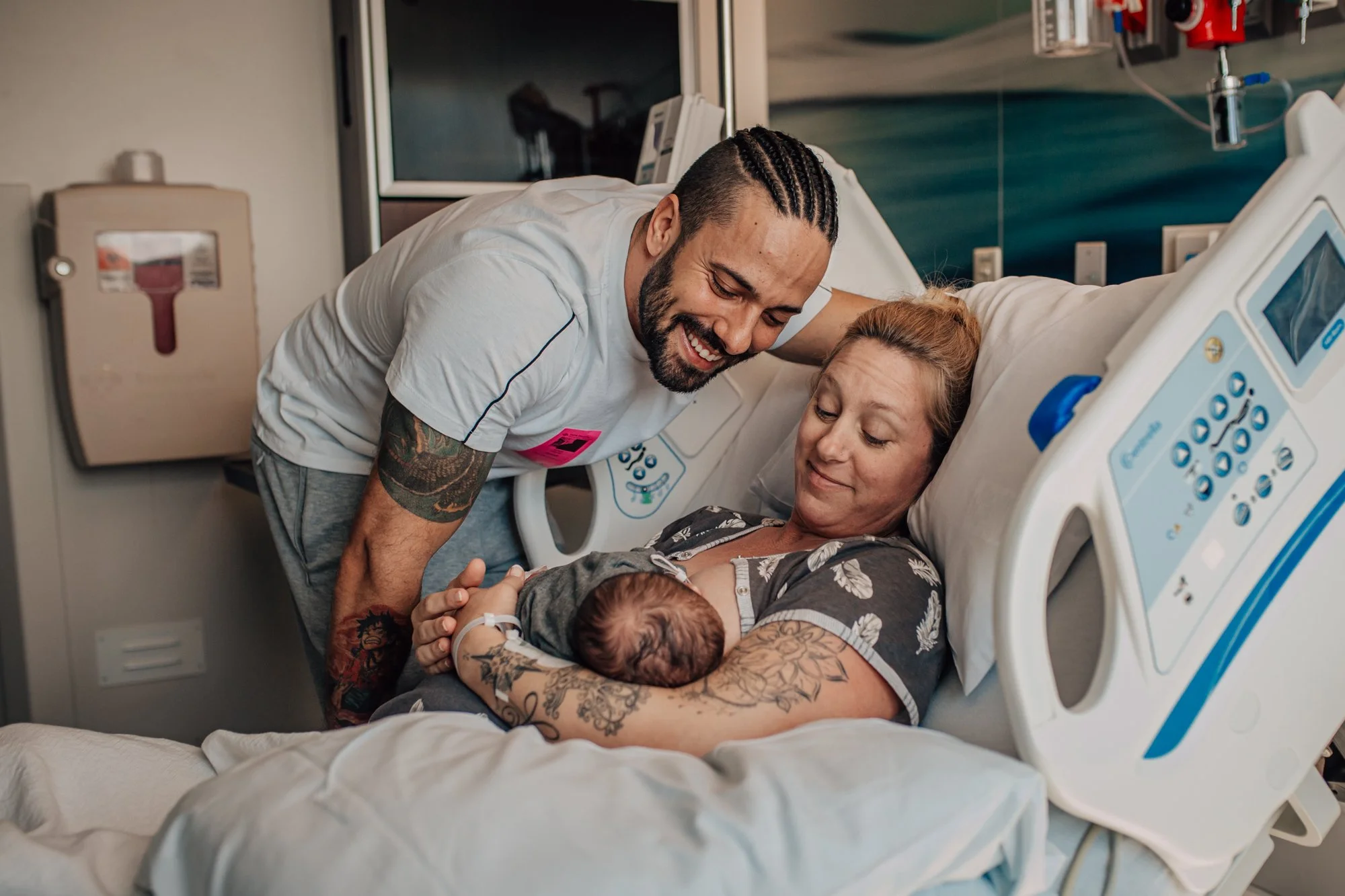 A man with braided hair and tattoos is laughing and leaning over a hospital bed, holding hands with a woman lying in the bed who is smiling, with a newborn baby on her chest in a hospital room.
