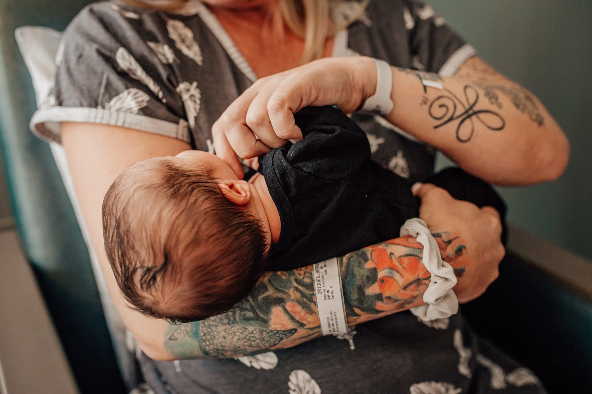 A person holding a newborn baby in their arms. The person has tattoos on their arm, a wristband, and is wearing a dark shirt with a pattern. The baby is dressed in black and is resting its head on the person’s arm.
