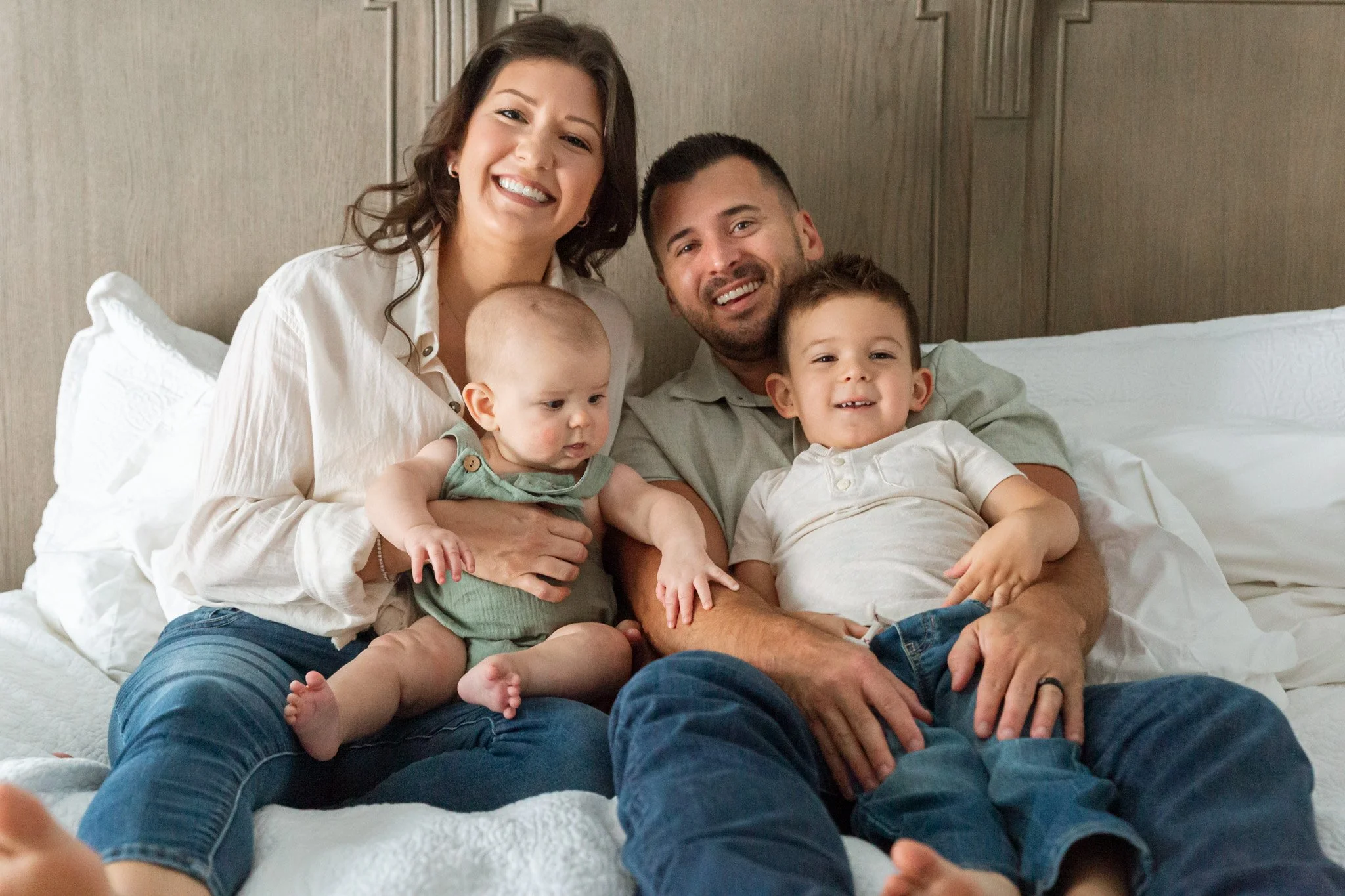 A family of four smiling and sitting together on a bed, with a wooden headboard in the background. The mother, father, and two young boys are visible, all looking happy.