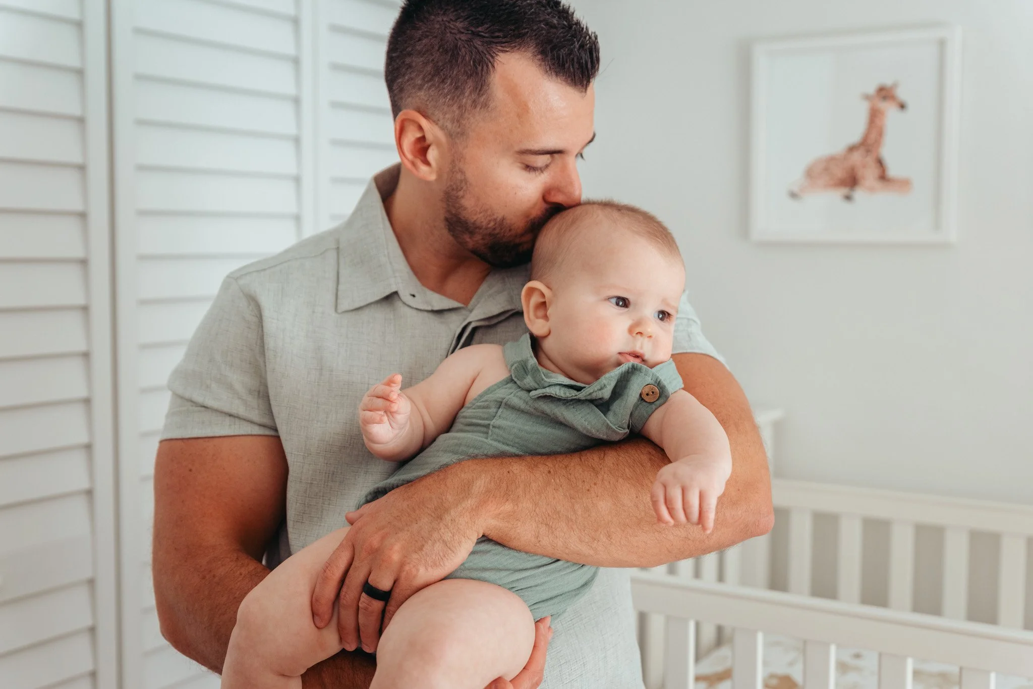 A man with short dark hair and a beard gently kisses a baby on the forehead while holding the baby in his arms. The baby has light skin, light hair, and is wearing a green sleeveless outfit. The background features white slatted walls and a framed pi