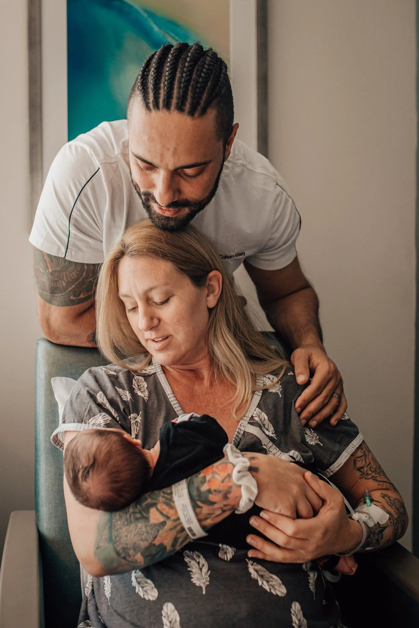 A woman holding a newborn baby with a man standing behind her, leaning in to look at the baby, inside a hospital room.