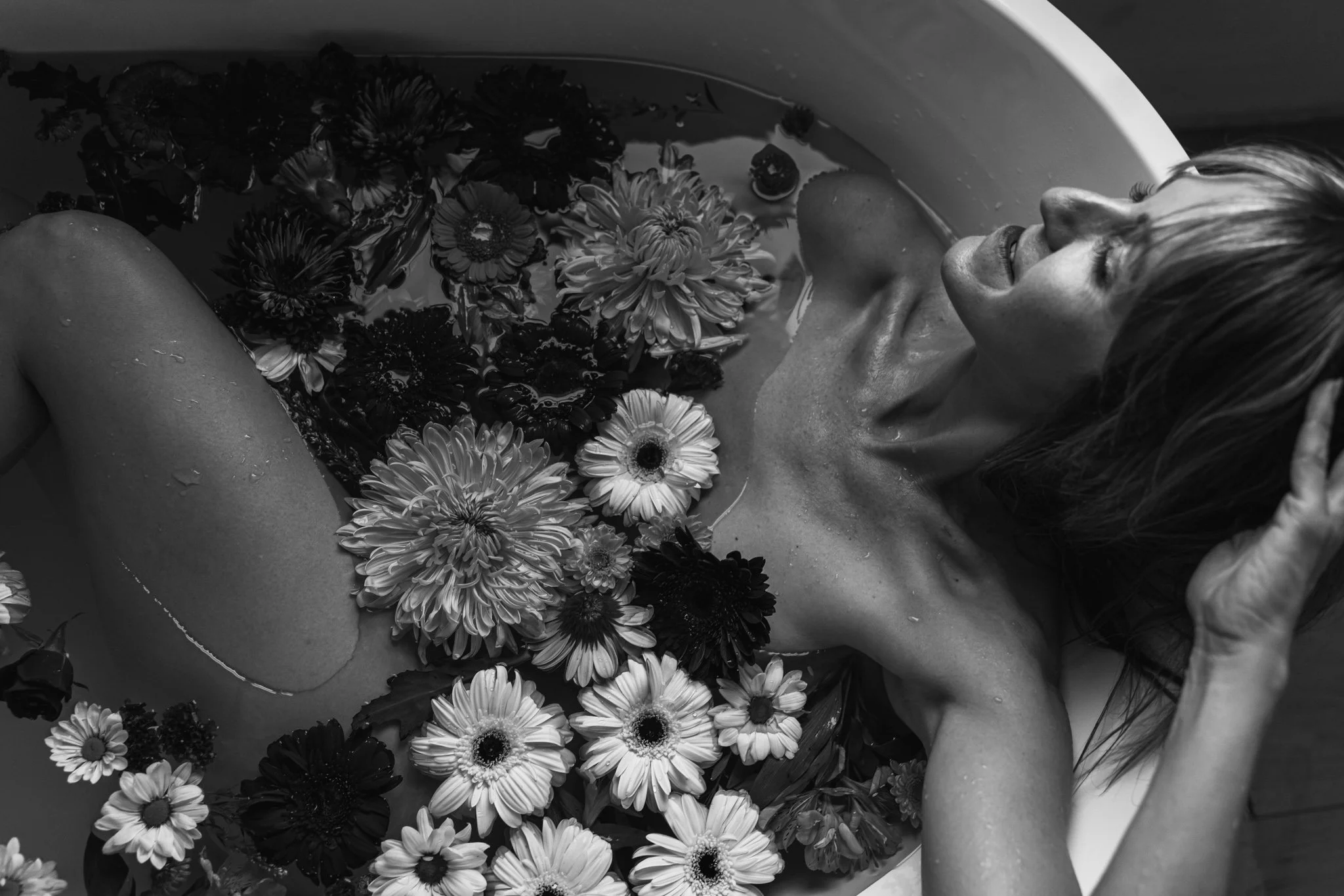 A woman with wet hair submerged in a bathtub filled with water and floating flowers, smiling with her eyes closed.