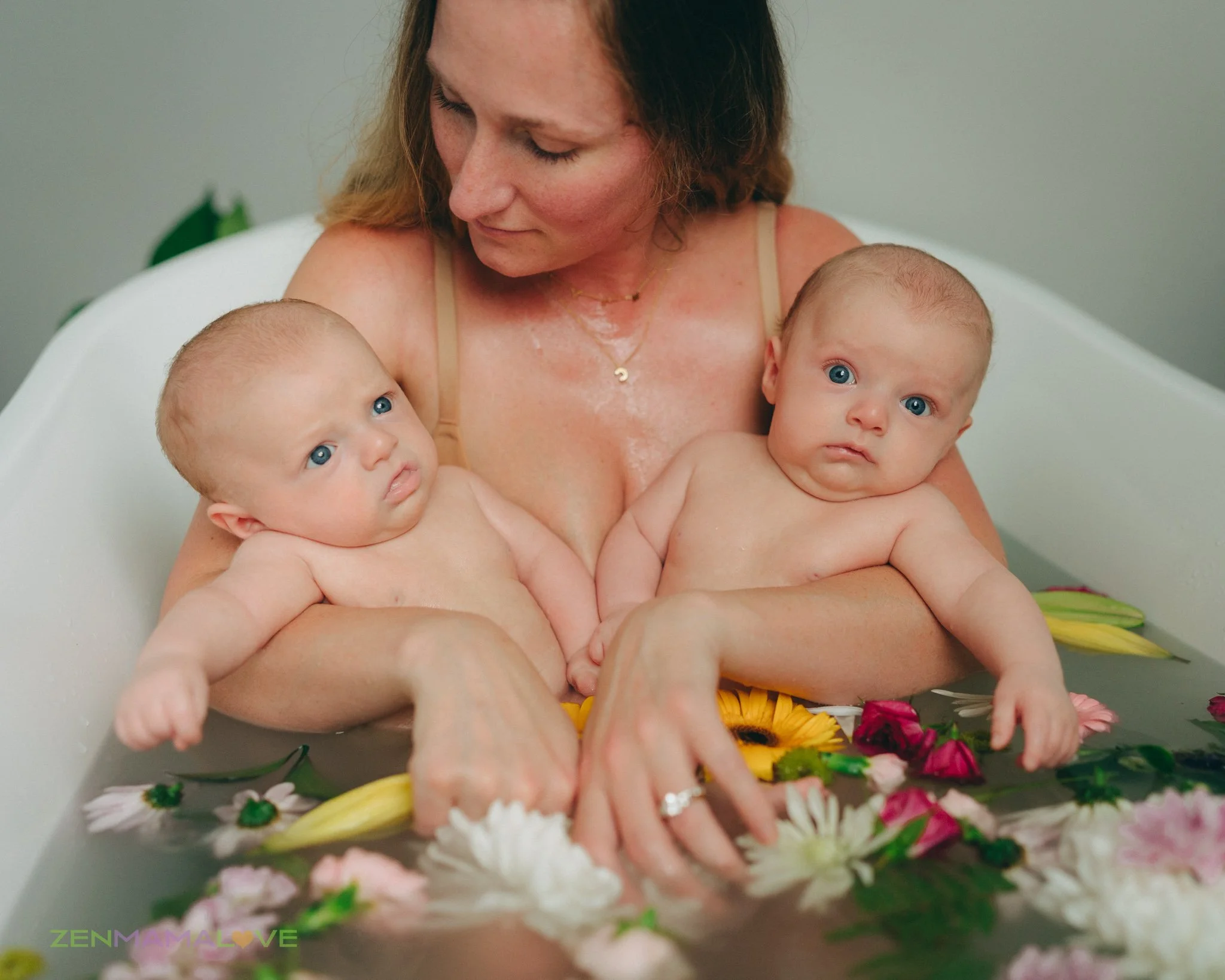 A woman with shoulder-length hair holding two babies with blue eyes, all in a bathtub filled with colorful flowers, looking at each other and the camera.