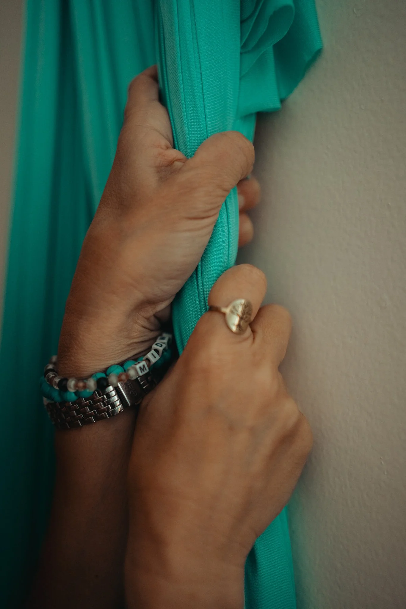 Close-up of a person holding a teal-colored fabric or object against a beige wall, wearing a beaded bracelet, a metallic watch, and a ring.