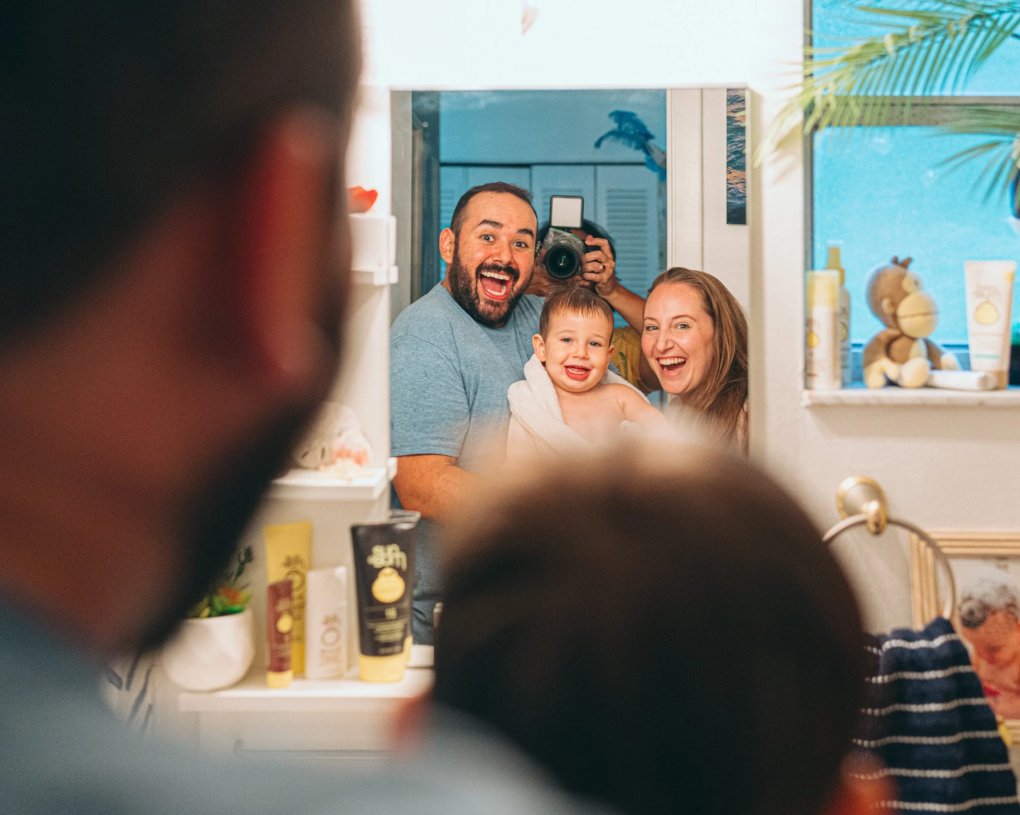 A smiling family of three is seen through a bathroom mirror, with an open medicine cabinet in the foreground. The family includes a man with a beard, a woman, and a young child. The bathroom has a blue color scheme with beach-themed decor, and variou