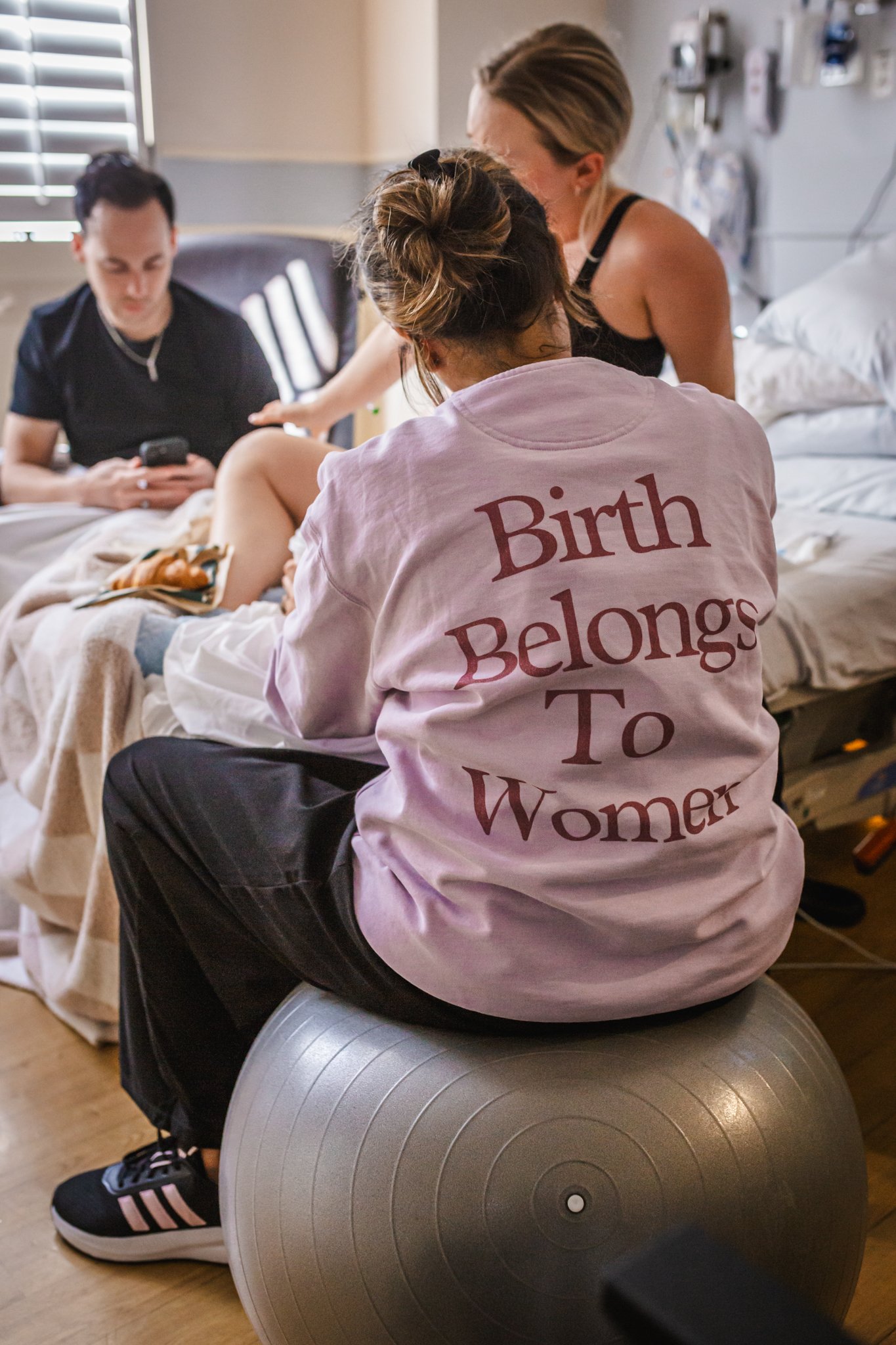 A woman with her hair in a bun, wearing a pink shirt that says 'Birth Belongs To Women,' sitting on a silver exercise ball in a hospital room. She is talking to a woman standing beside her and a man sitting on a chair in the background looking at a p