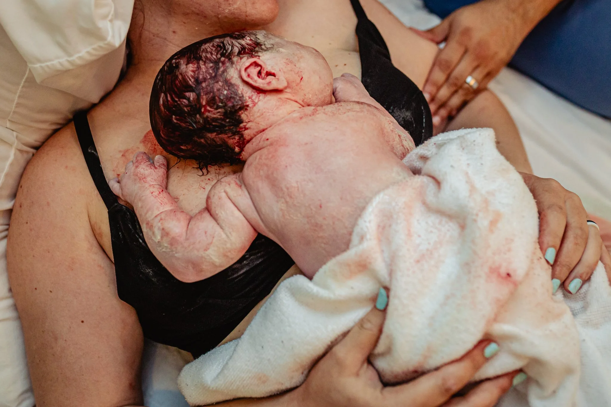 A newborn baby cradled in someone's arms during birth, with the baby's head still covered in blood and vernix, and the mother partially visible.