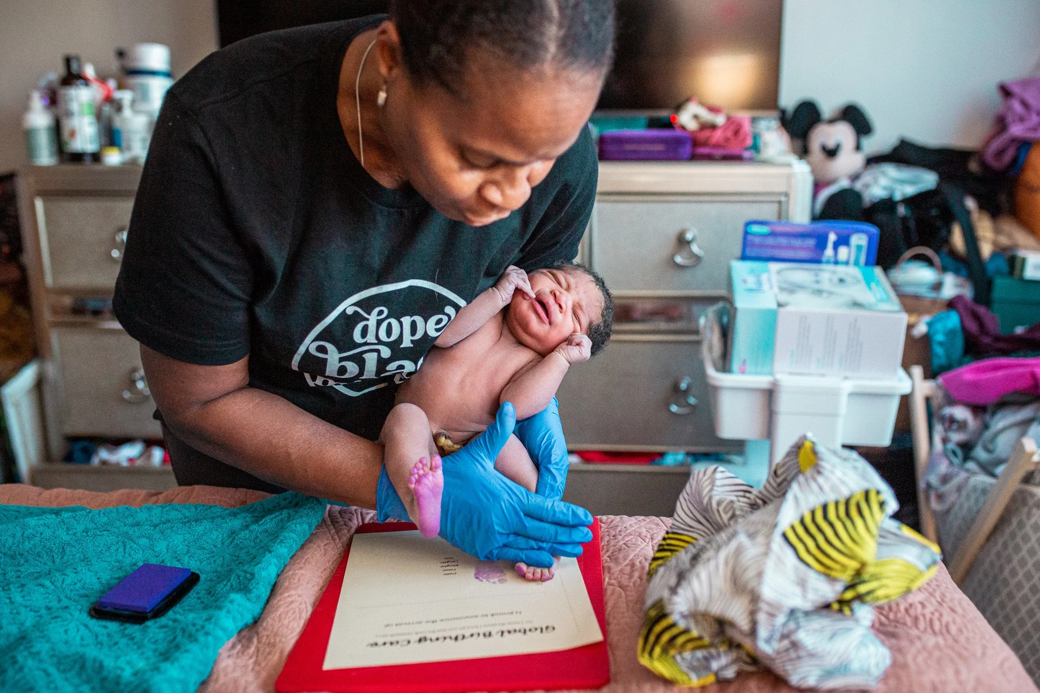 A woman looking down at a newborn baby she is holding in her arms, in a cluttered room with various items on a dresser and a table.