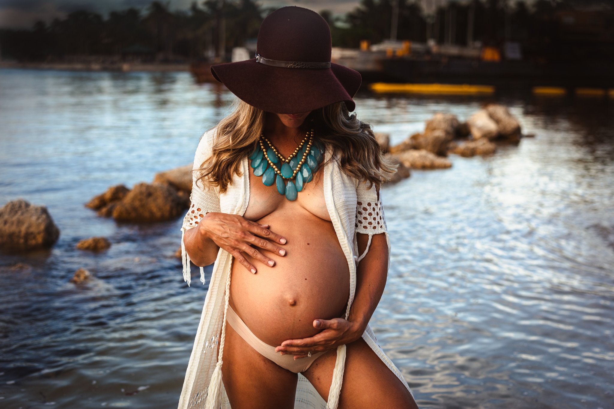 A pregnant woman standing by a river at sunset, wearing a large brown hat, a white open cardigan with lace details, and a chunky turquoise necklace, gently touching her belly with both hands.