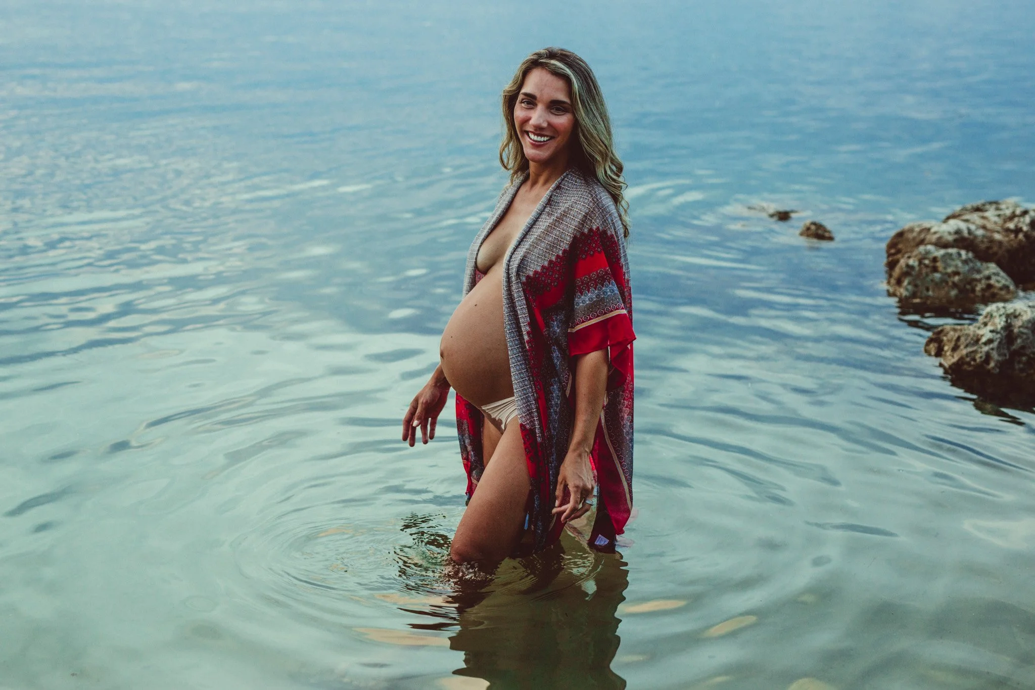 A pregnant woman standing knee-deep in water by the rocks of a shoreline, smiling and looking at the camera during daytime.