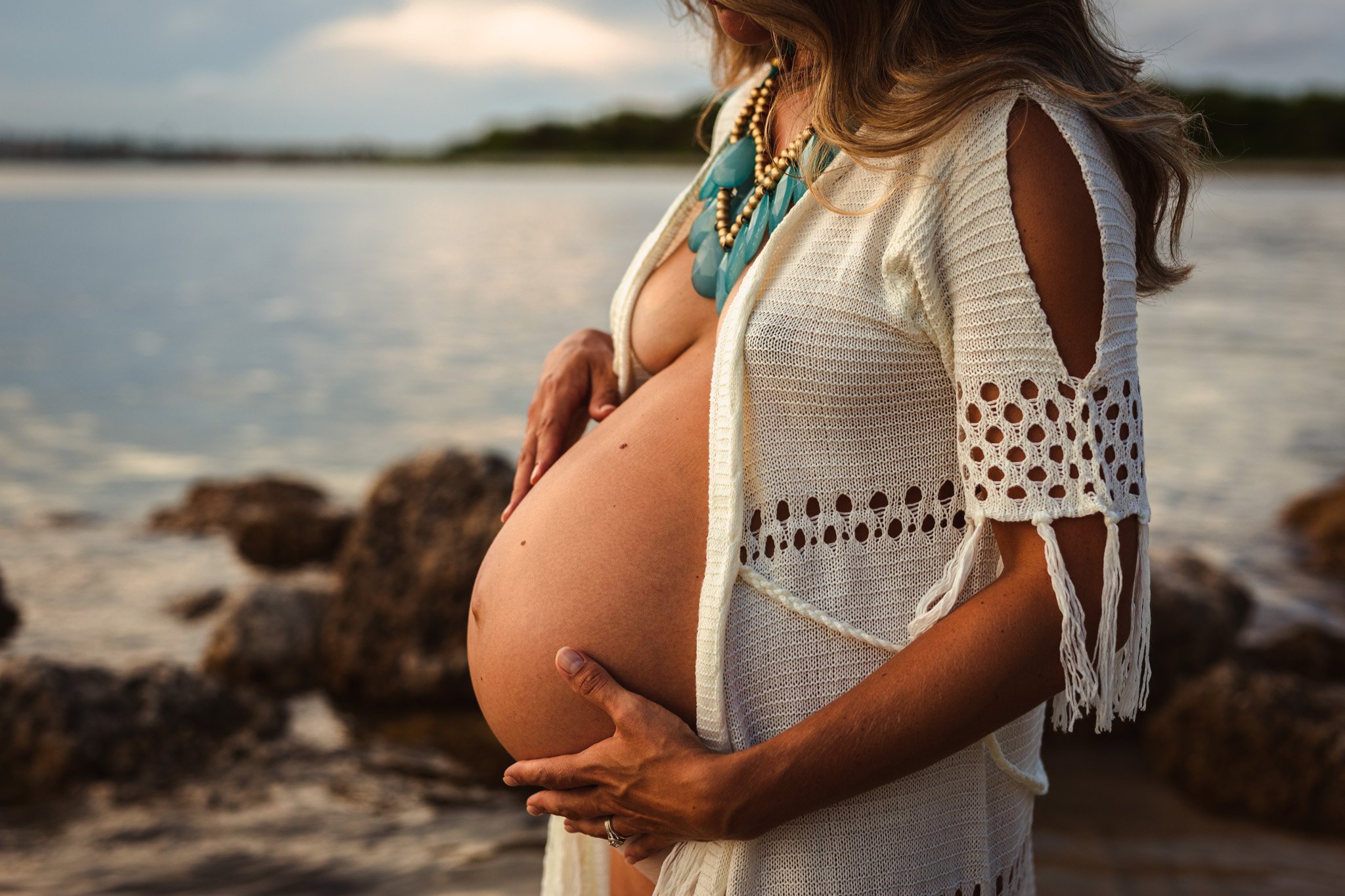 A pregnant woman with long hair wearing a white, crocheted cover-up and multiple blue and beige necklaces, standing near water with rocks in the background, and holding her belly with both hands.