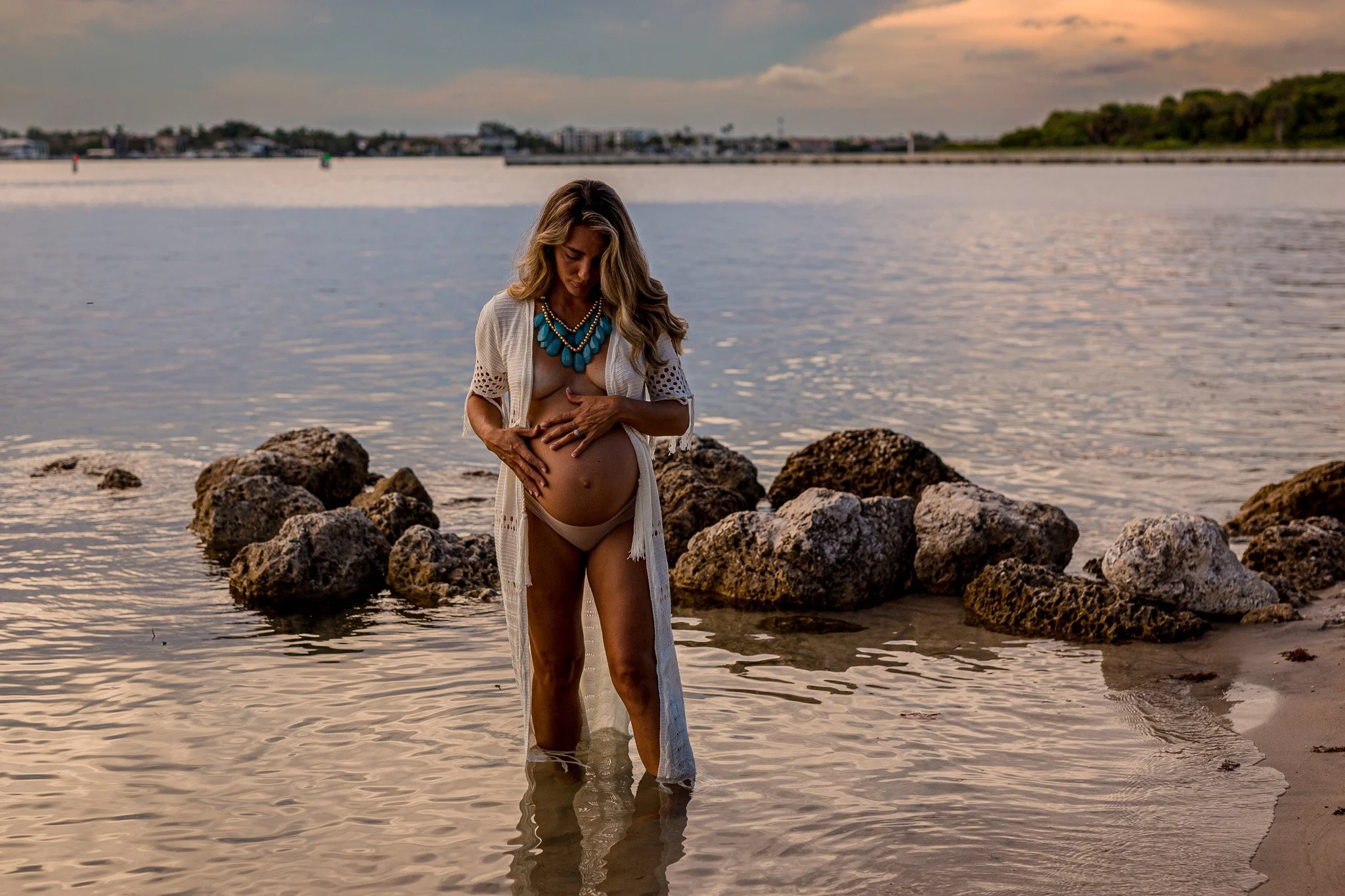 Woman standing in water near rocks at the beach during sunset, wearing a beige bra, a white cover-up, and a turquoise necklace.