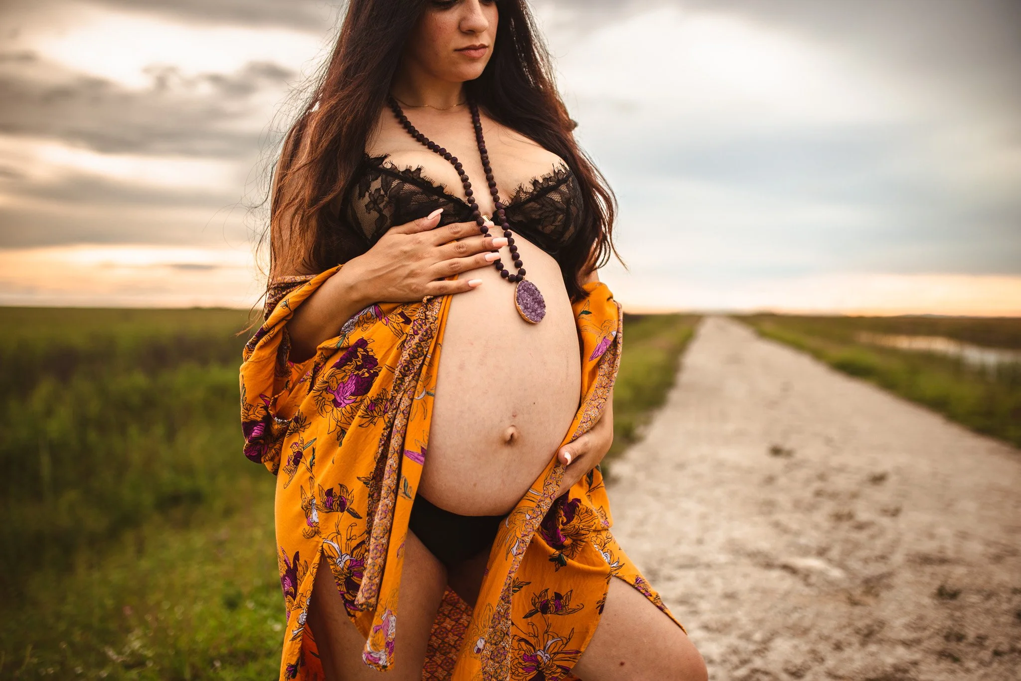 A woman with long dark hair wearing black lace lingerie and an orange floral robe, holding her pregnant belly with a spiritual-looking necklace, standing outdoors on a dirt path with a cloudy sky in the background.