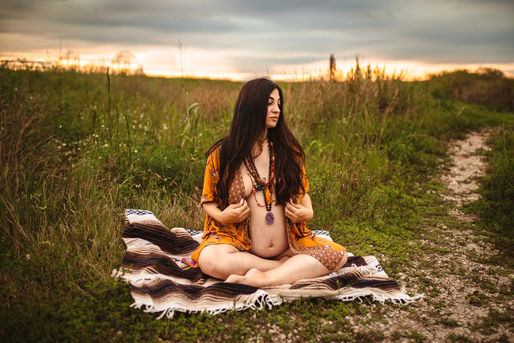 A pregnant woman sitting cross-legged on a blanket outdoors at sunset, wearing colorful jewelry and a loose yellow top, with eyes closed.