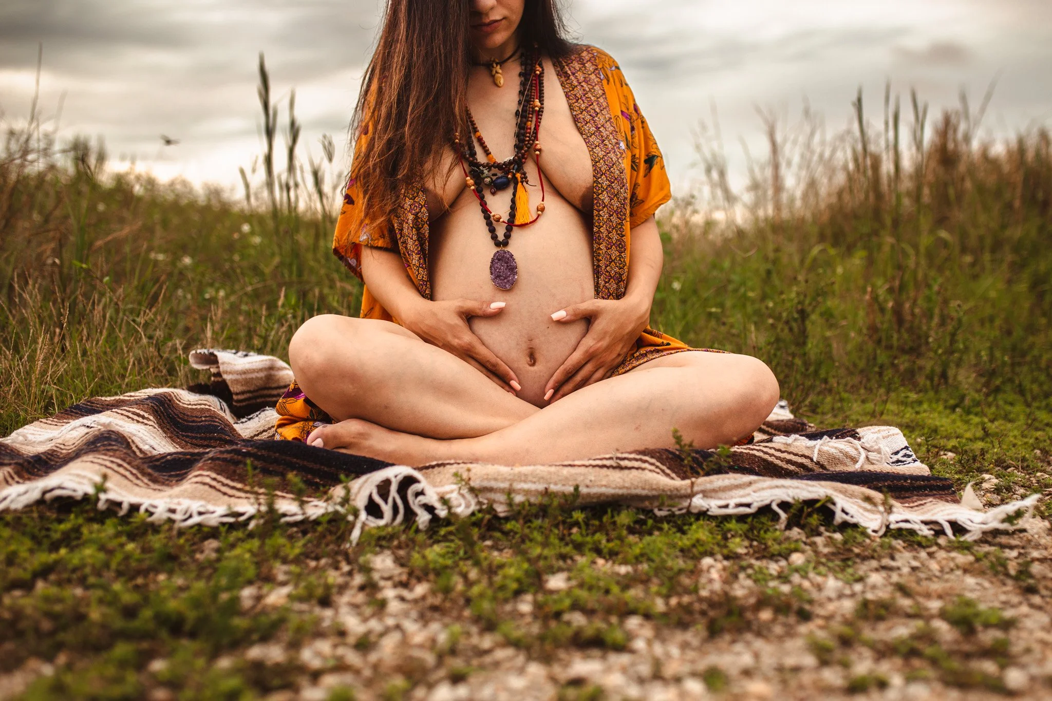 A woman sits cross-legged on a patterned blanket in an outdoor natural setting, with a cloudy sky overhead. She wears multiple layered necklaces and an open, colorful kimono-style cover-up, exposing her bare chest and abdomen. Her long, dark hair cas