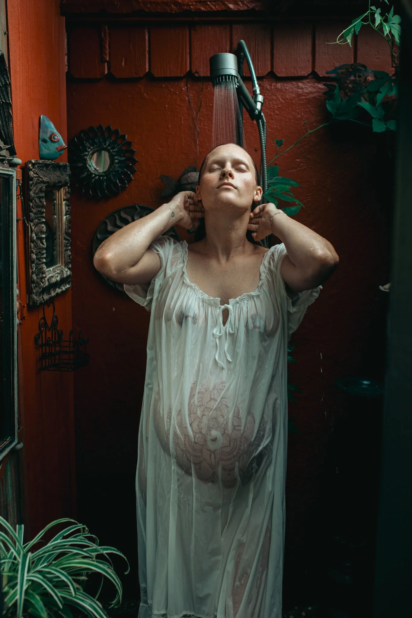 A person standing in a shower with water spraying down, eyes closed, wearing a wet, semi-transparent white dress, in a cozy outdoor space with red wooden walls, green plants, and decorative items.