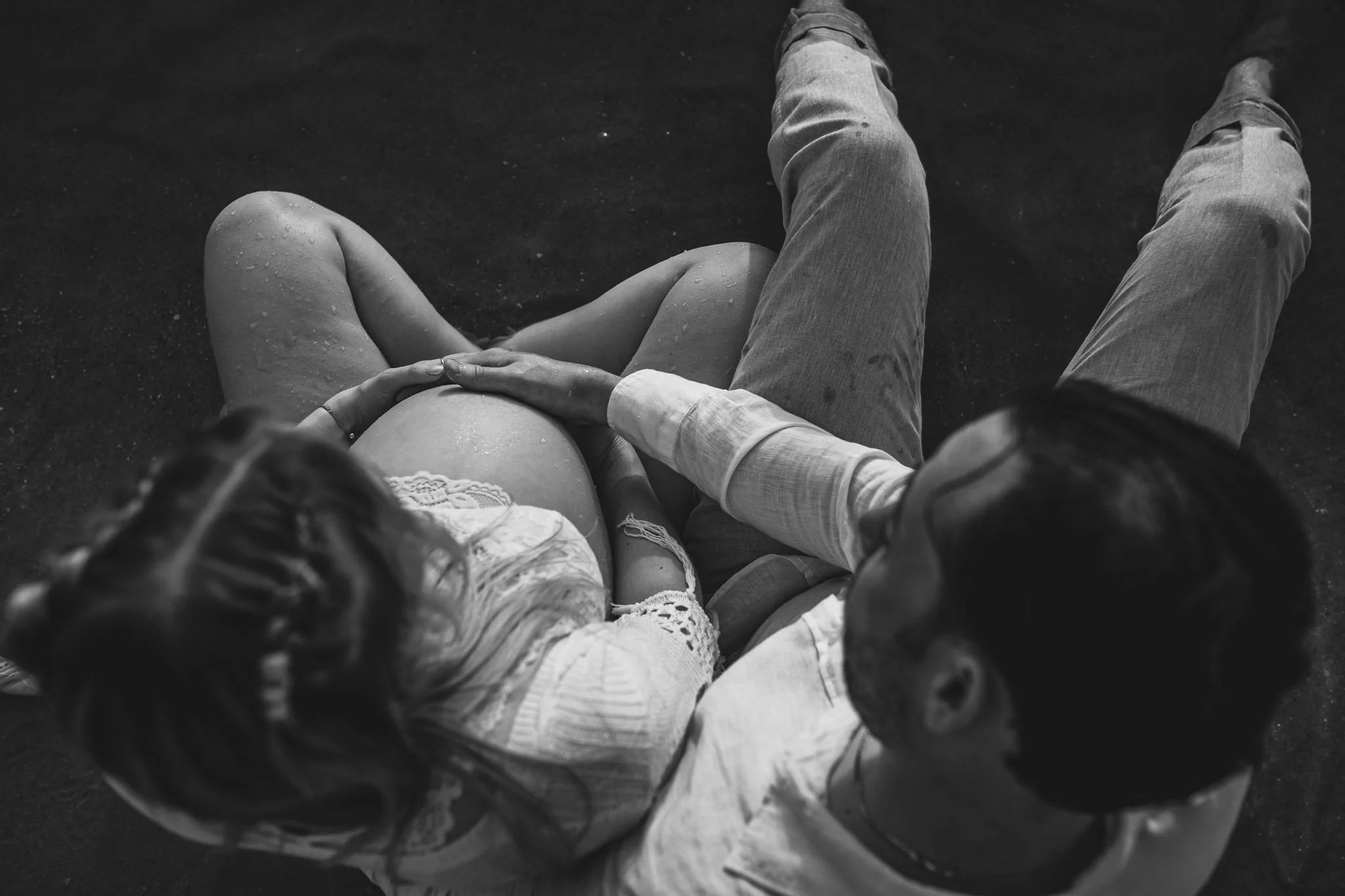 A black and white photo of a person sitting on a dark surface, with their legs stretched out in front. They are wearing light-colored jeans and a long-sleeve shirt, and holding hands with a child who is sitting beside them. The child is wearing lace-