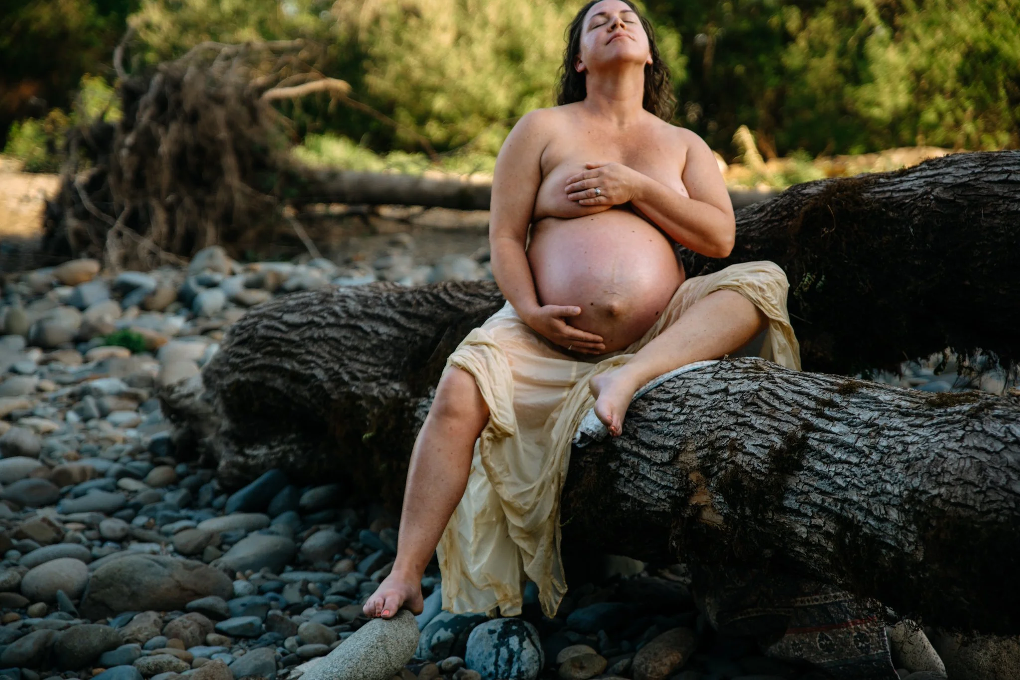 A woman with dark hair, sitting topless on a fallen tree trunk outdoors, with her eyes closed and hands covering her chest. She is wearing a light-colored skirt, and her pregnant belly is visible. The background features green foliage and a rocky, pe