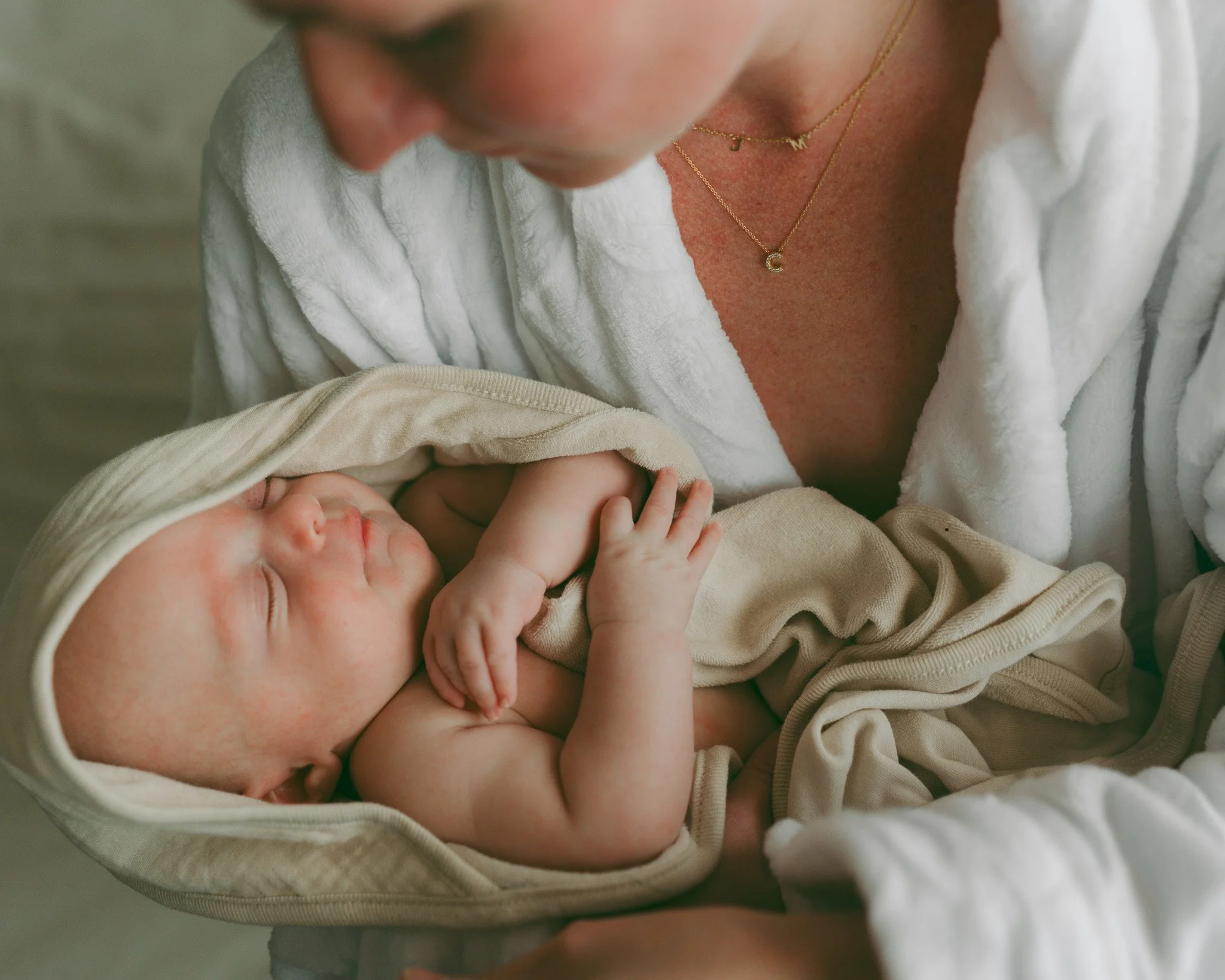 A close-up of a woman holding a sleeping baby, with the woman wearing layered gold necklaces with 'J', 'M', and a crescent moon pendants, dressed in a white textured shirt, and the baby wrapped in a beige blanket with a light-colored bib.