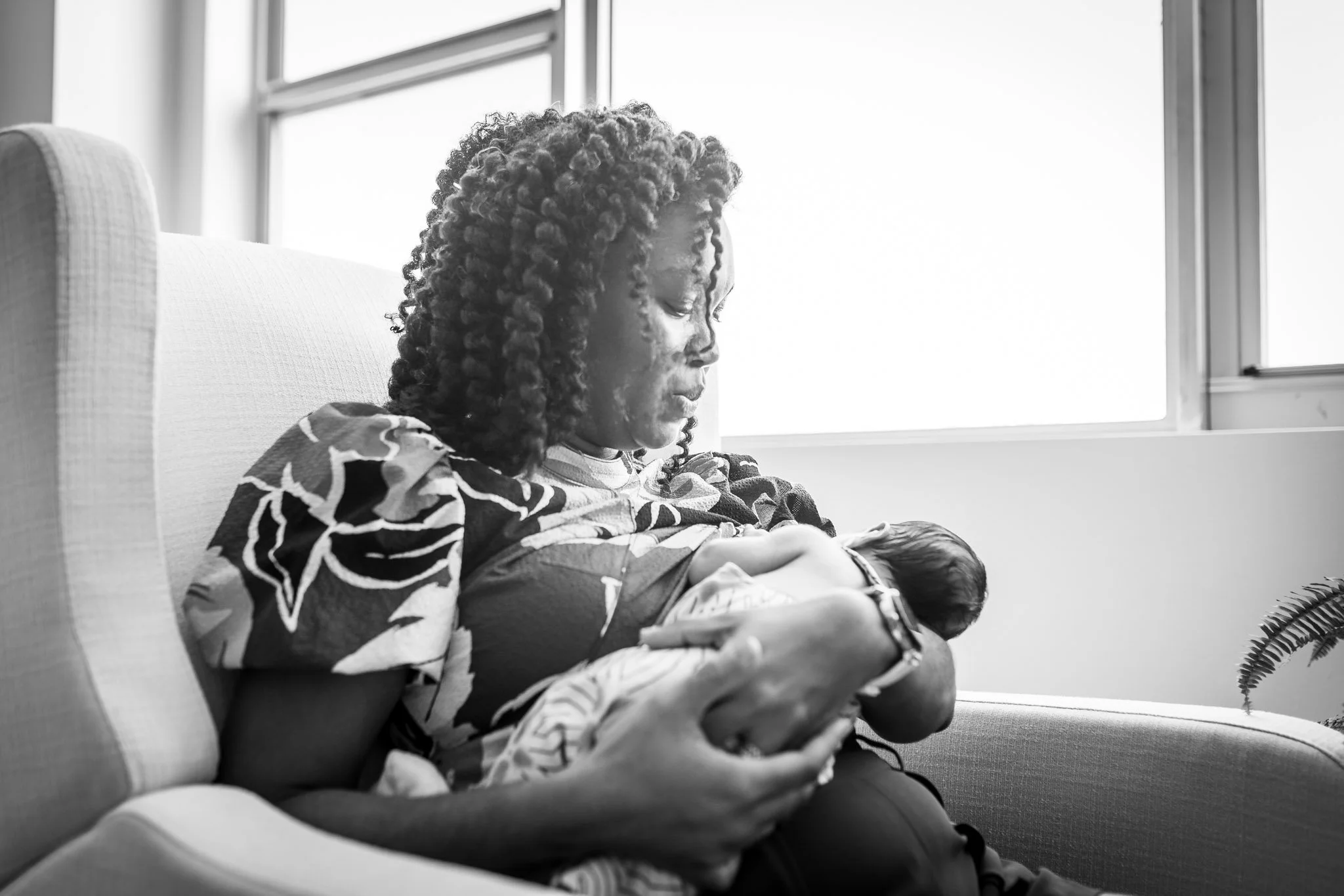 A woman with curly hair sitting on a couch, holding a baby in her arms near a window.