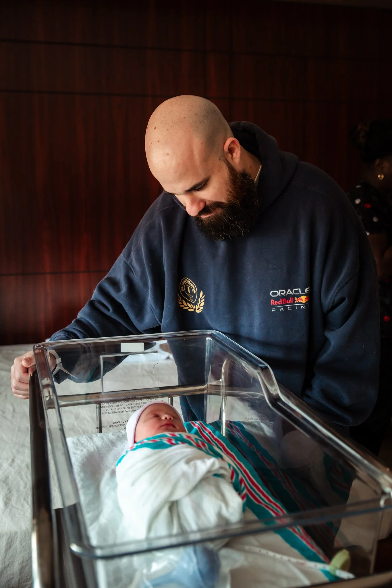 A man with a beard and a shaved head wearing a dark hoodie looks down at a newborn baby in an incubator.