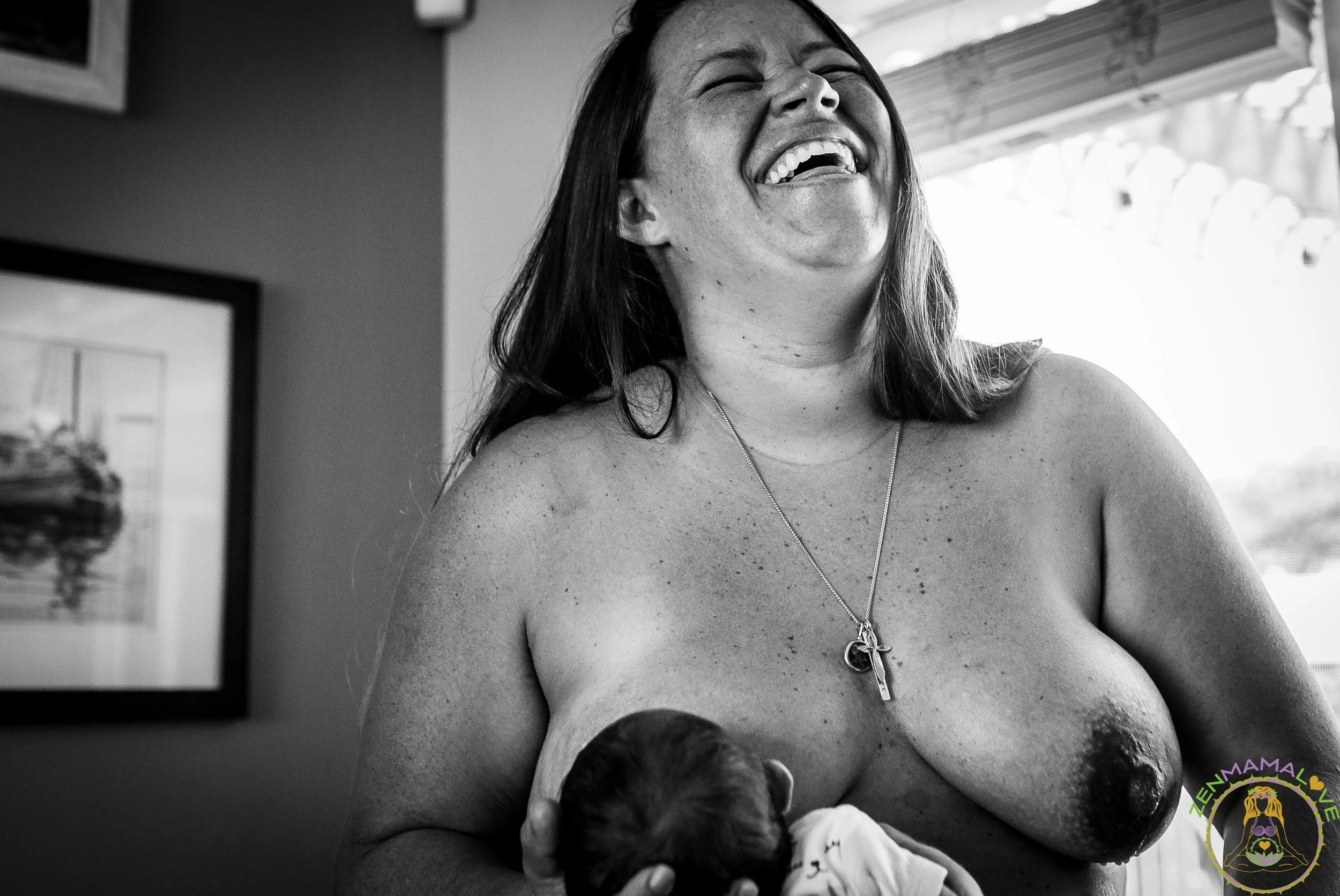 A woman laughing while breastfeeding a baby, sitting indoors with a window in the background.
