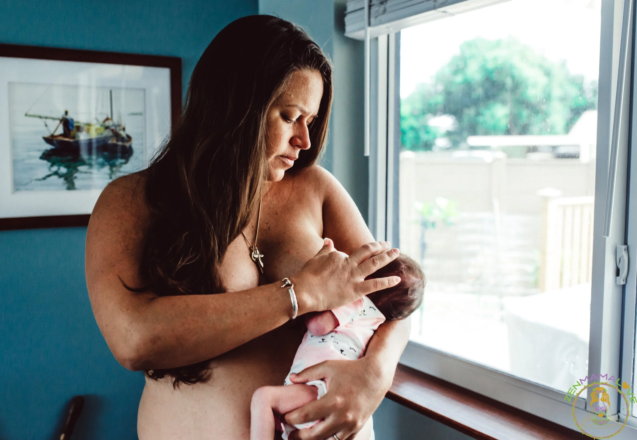 A woman holding a newborn baby near a window in a room with blue walls, with a framed picture of a boat on water hanging on the wall.