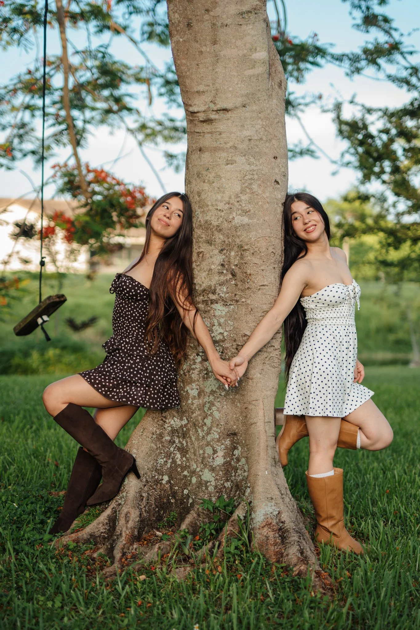 Two women in vintage dresses holding hands and leaning against opposite sides of a large tree in a grassy park, with trees and a swing in the background.