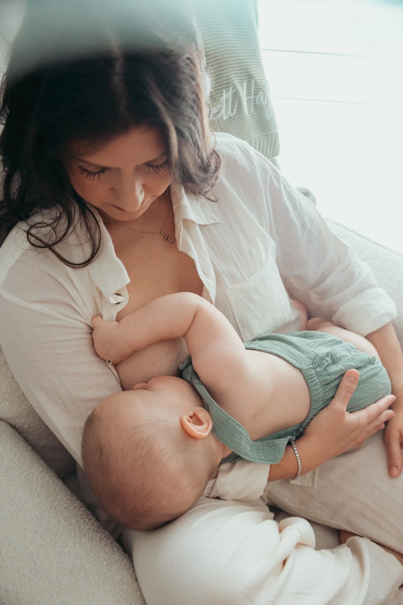 A woman sitting on the floor, breastfeeding a toddler with both crying babies lying on her lap, in a cozy, well-lit room.
