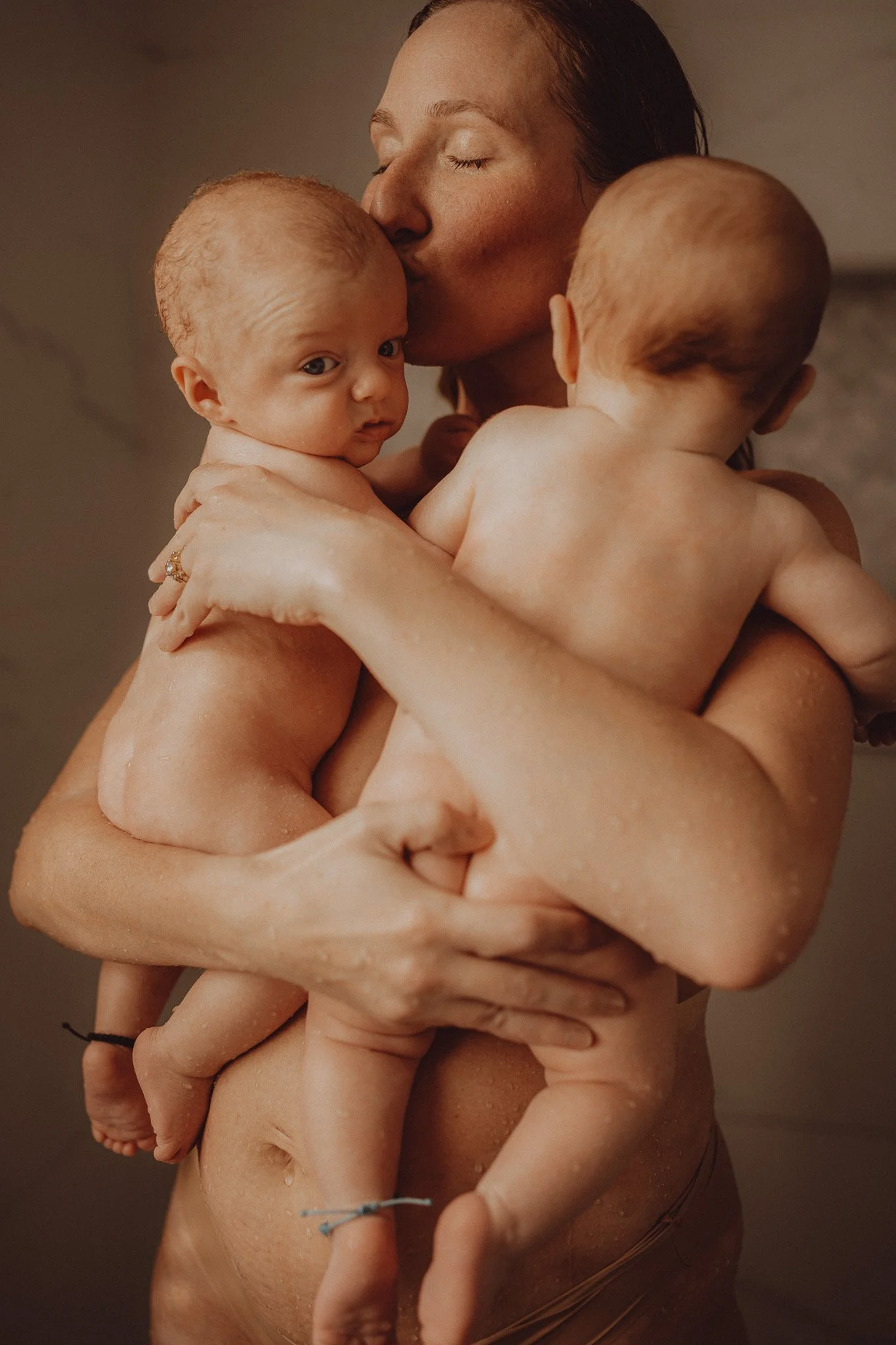 A woman holding two babies, one resting on her chest and the other on her shoulder, with a warm, intimate moment of affection and closeness.